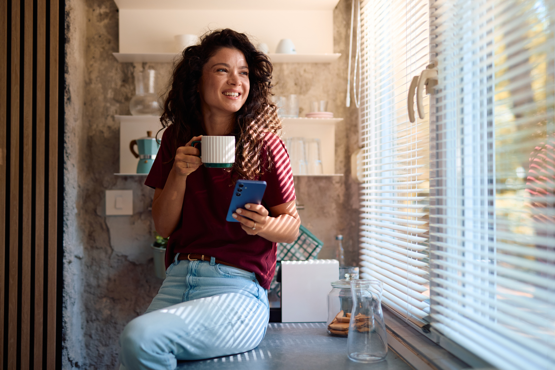 A woman with curly hair smiling while holding a mug and looking out of a window with blinds in a cozy kitchen or dining area.