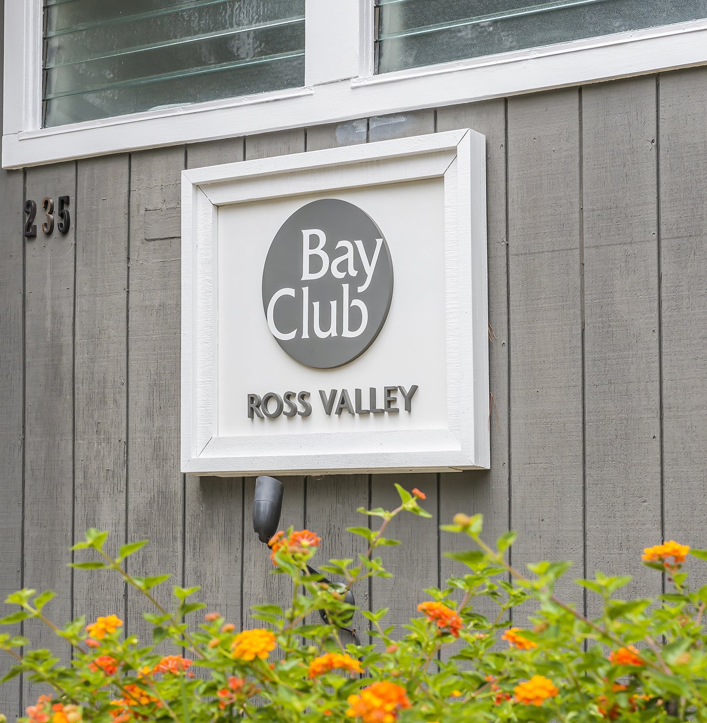 Sign for Bay Club Ross Valley mounted on a gray wooden wall, with orange flowers at the bottom of the image.