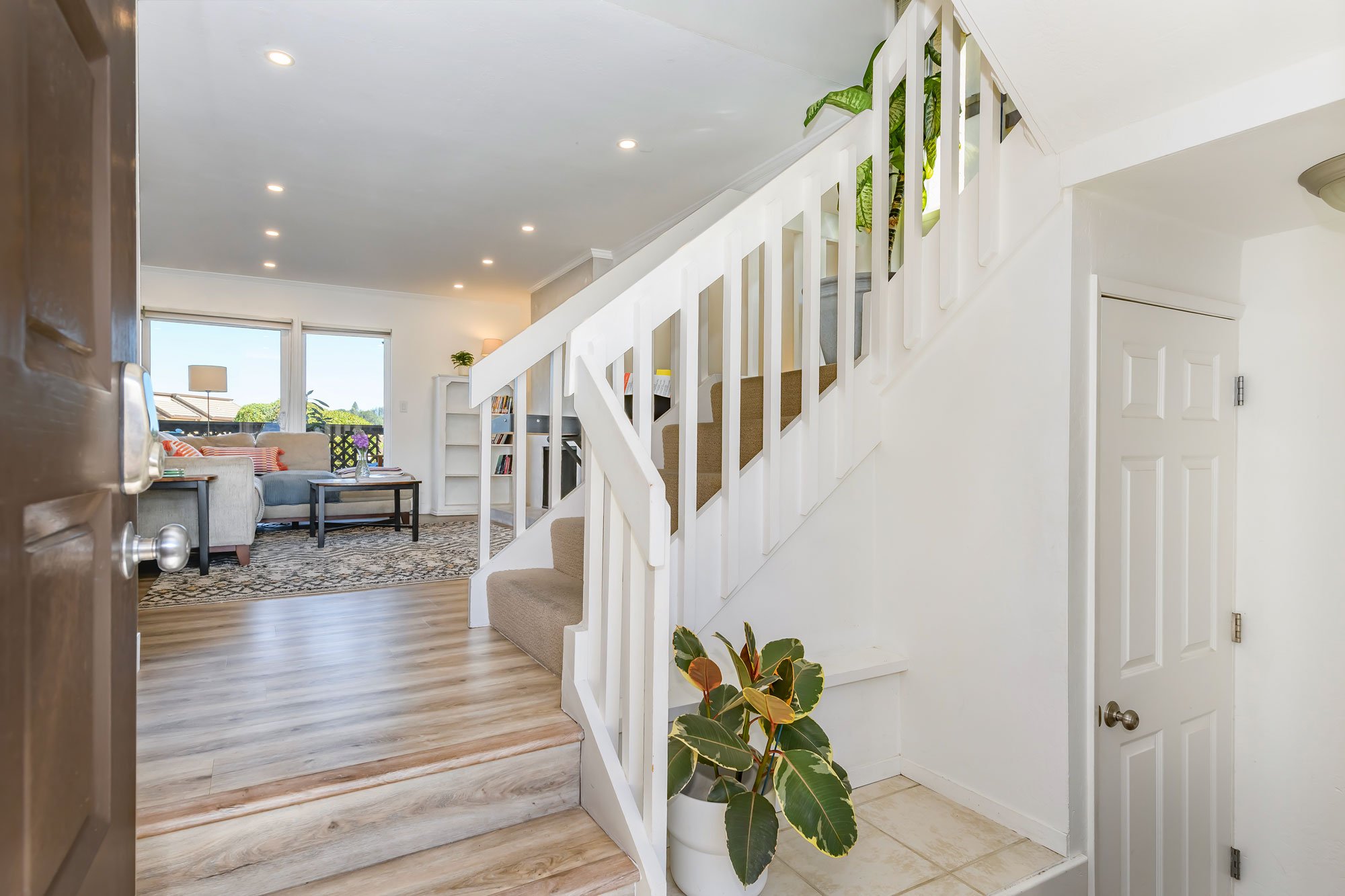 Entryway with stairs, potted plant, and view into living room with sofa and large windows.