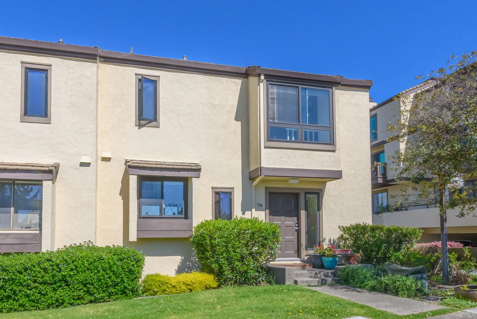 Front view of a beige multi-family residential building with brown window frames and a brown door, surrounded by green bushes, plants, and a manicured lawn under a blue sky.