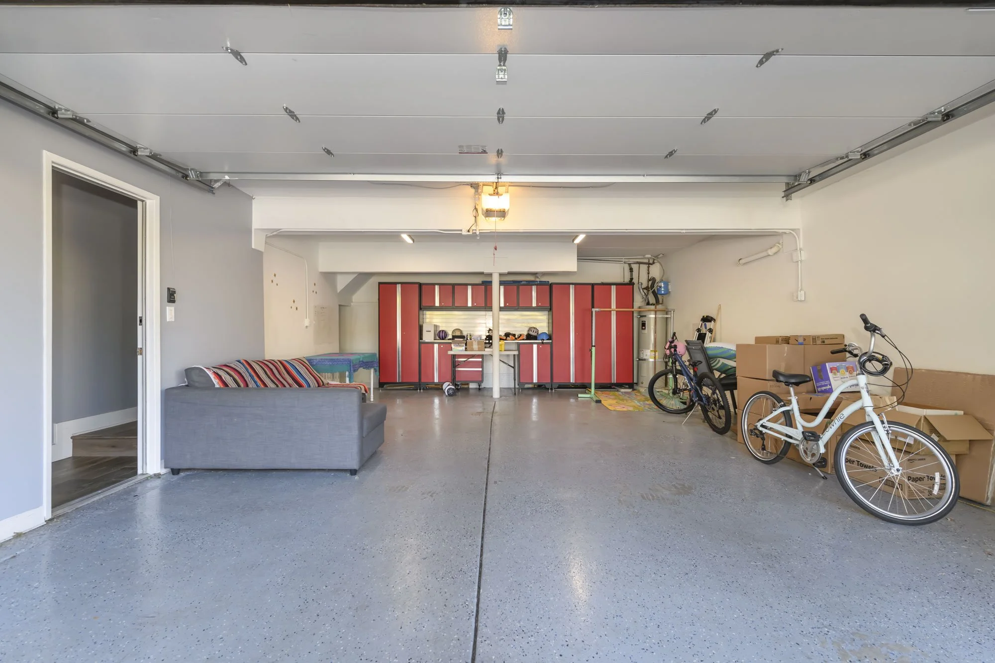 Garage with red storage cabinets, bicycles, boxes, and a gray couch on a polished concrete floor.