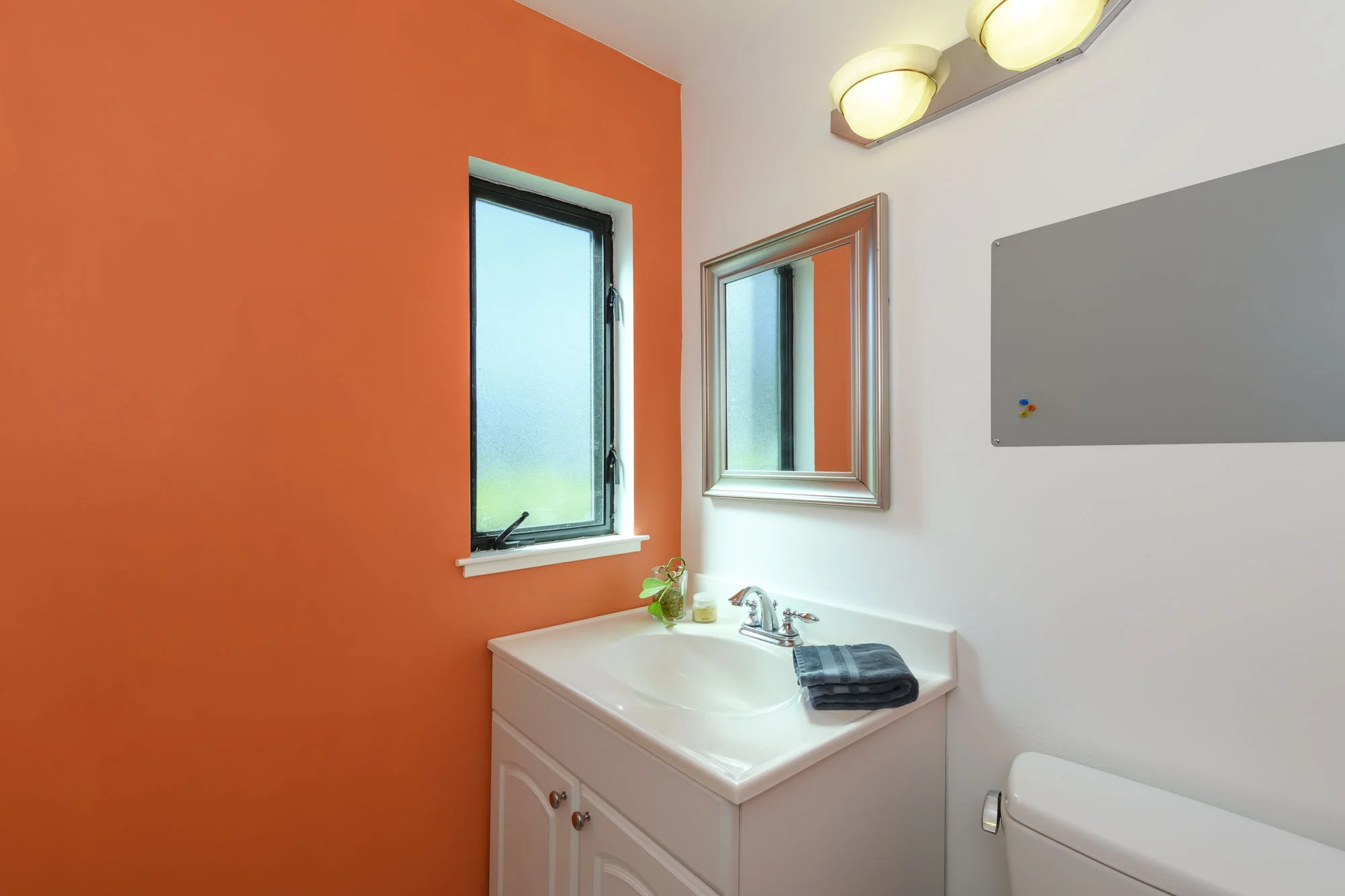 Bathroom with an orange accent wall, a small window, a mirror, a white vanity with a sink, a gray towel, and a toilet.