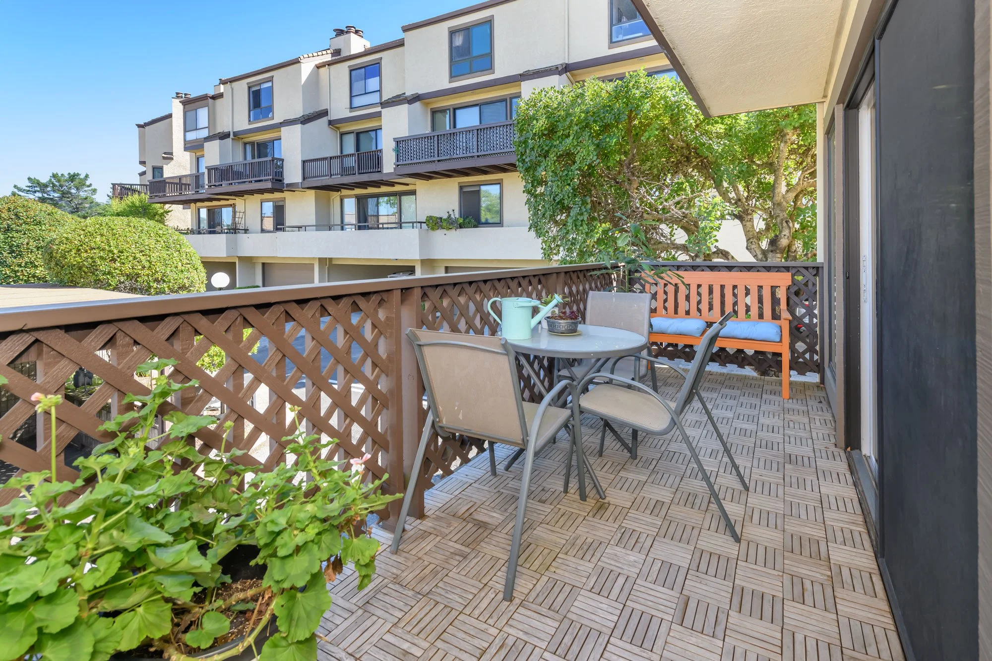 Balcony with wooden floor tiles, a table with beige chairs, a garden bench, potted plants, and a tree, overlooking neighboring apartment buildings.
