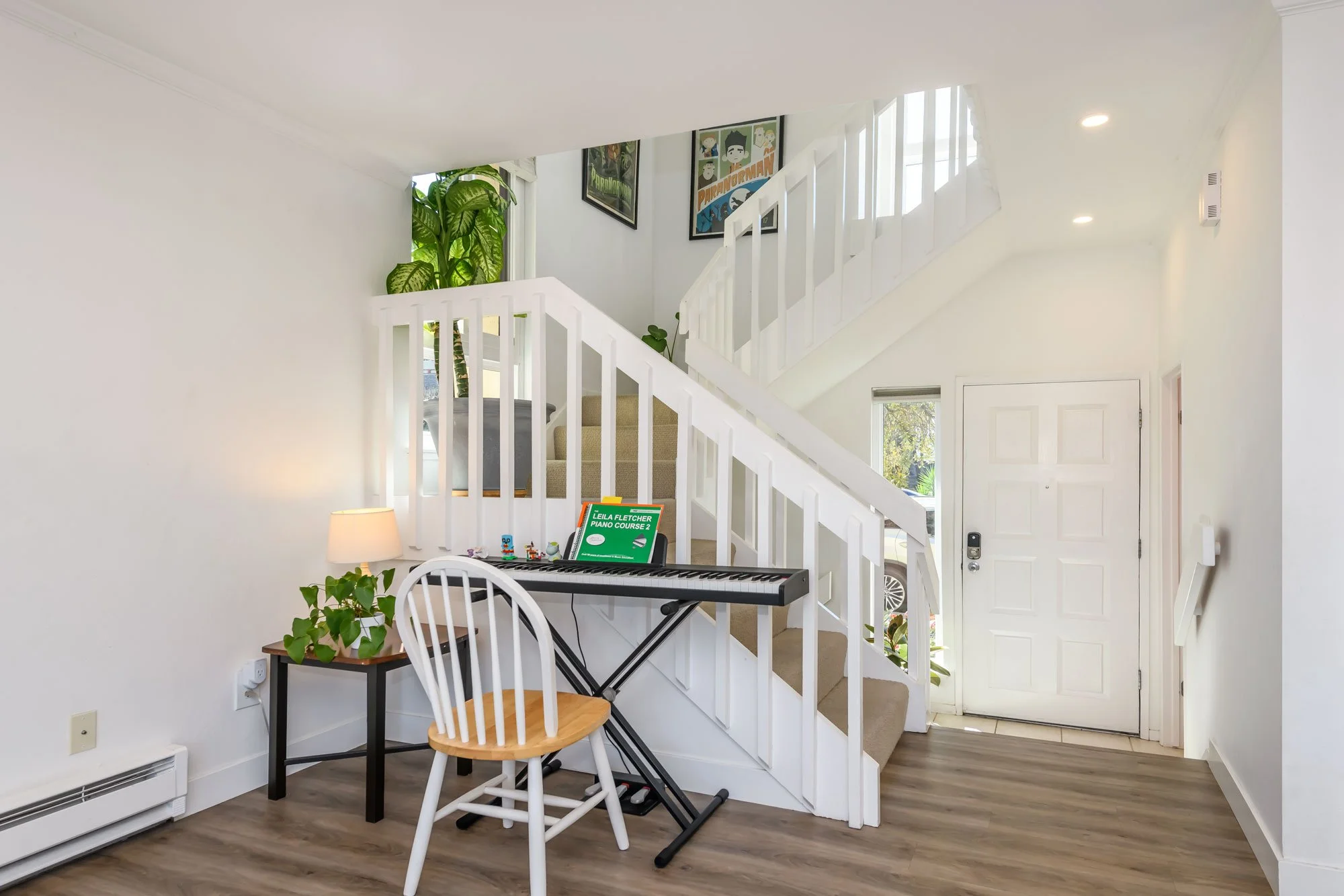 Interior view of a home with a staircase, a digital keyboard, potted plant, a table with a lamp, chairs, and framed artwork on white walls.