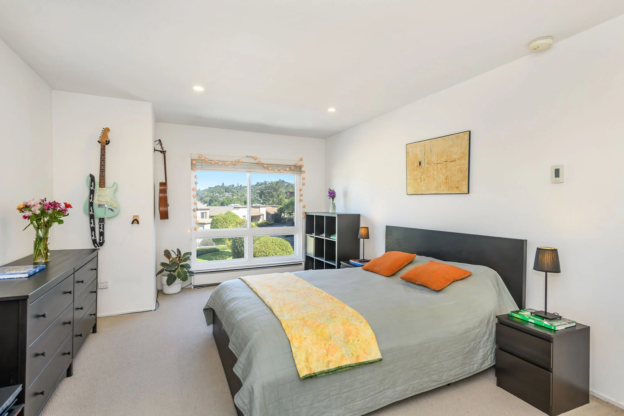 A bedroom with a bed, two black nightstands, a black bookshelf, a dresser, and a window with outdoor view and string lights