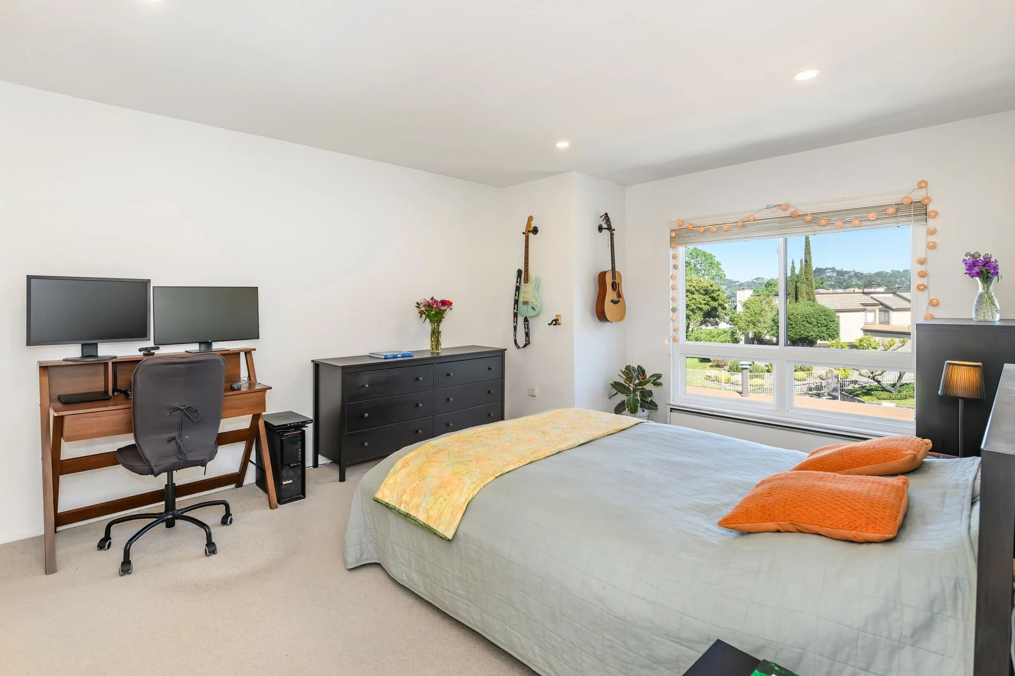 A bedroom with a bed, dresser, and workspace, with guitars on the wall and a window with a view of trees and houses outside.