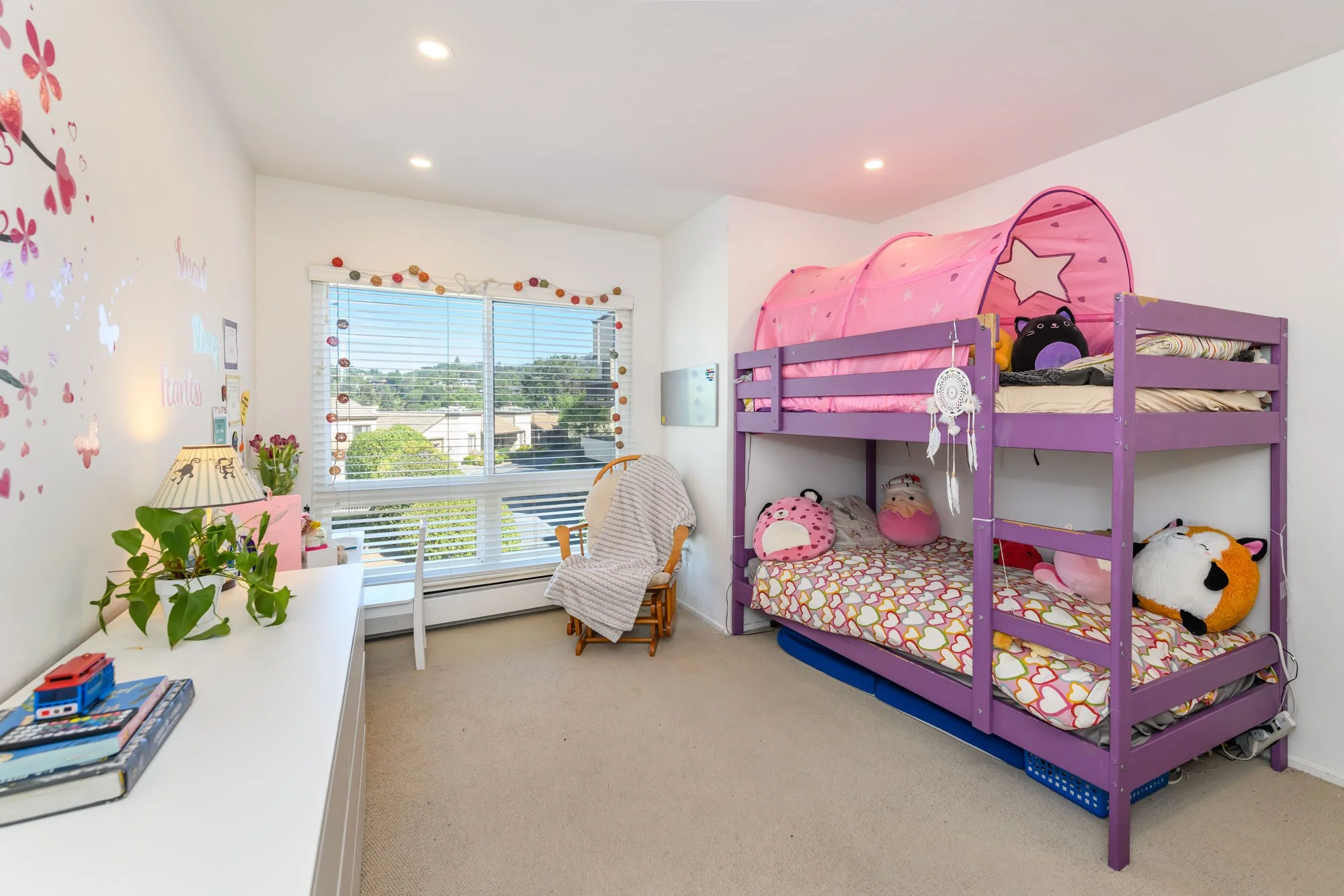 A children's bedroom with purple bunk beds decorated with plush toys, a white desk with books and a lamp, a window with blinds, and wall decor.