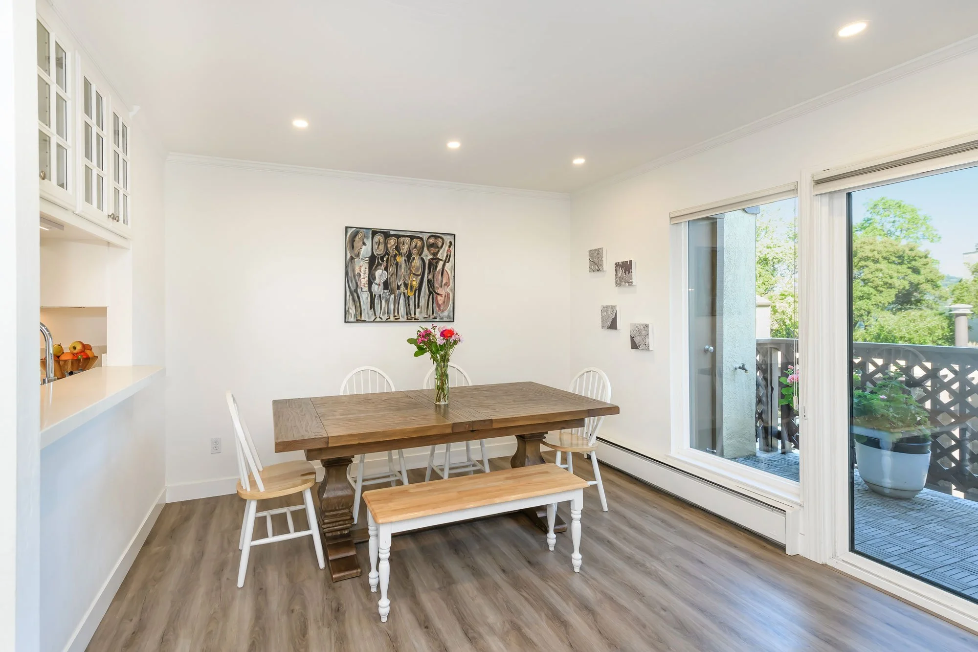 Dining room with wooden table, white chairs, and a bench, decorated with a vase of pink flowers; artwork on white walls; sliding glass door leading to a balcony with potted plants, overlooking trees.