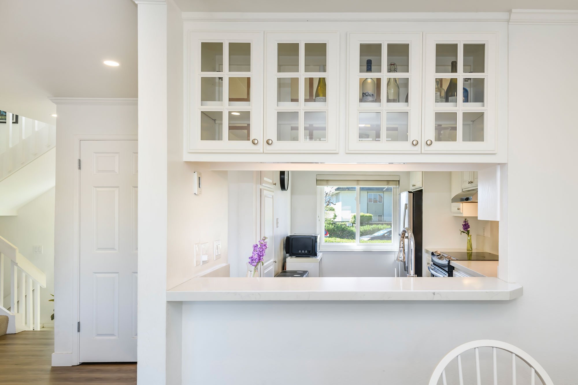 Modern white kitchen with upper glass-front cabinets, small black microwave on counter, window showing outdoor greenery, and purple flowers on counter.