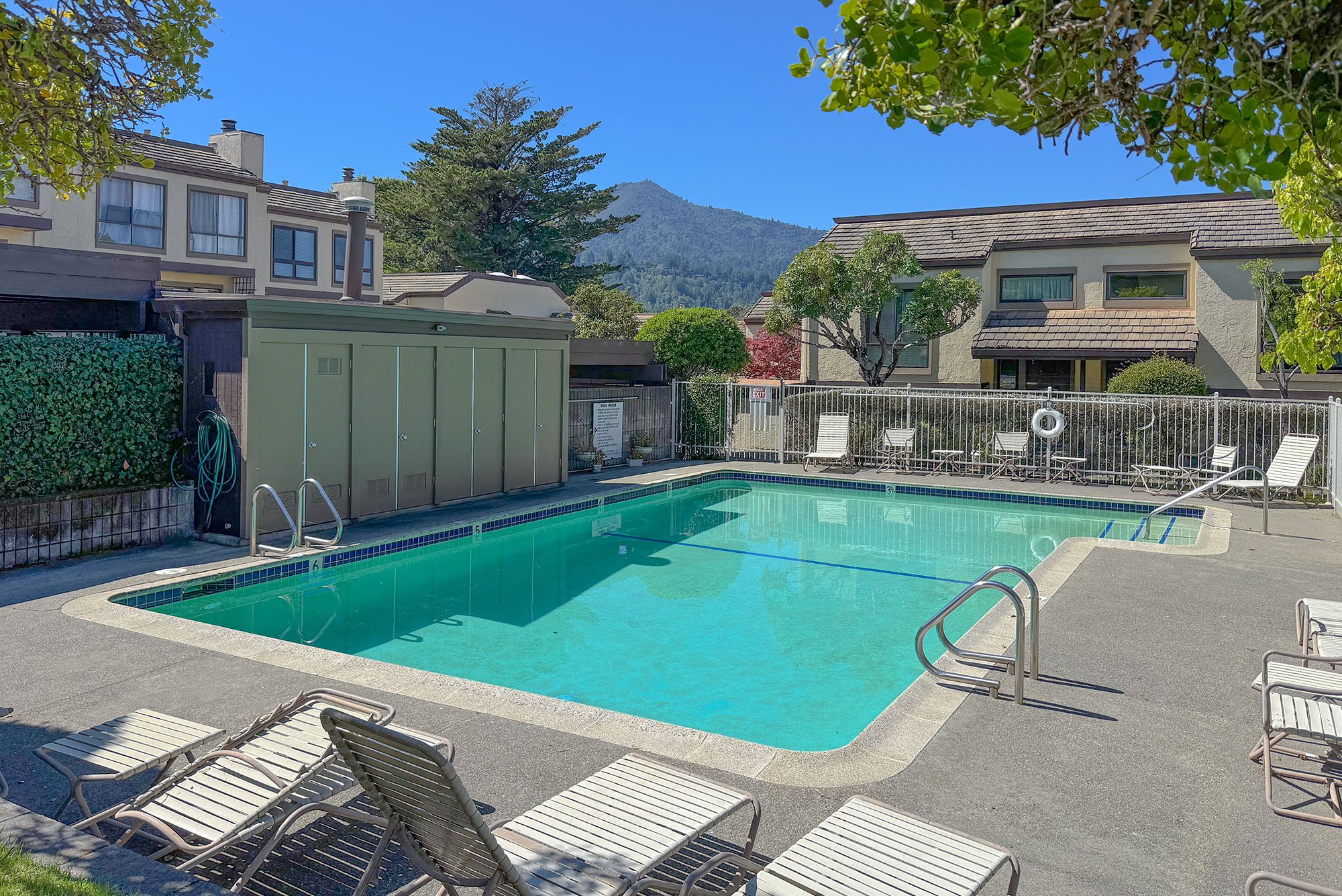 Outdoor swimming pool with a concrete deck, surrounded by lounge chairs and a safety fence. Nearby, there is a storage shed, residential buildings, trees, and a mountain in the background under a clear blue sky.