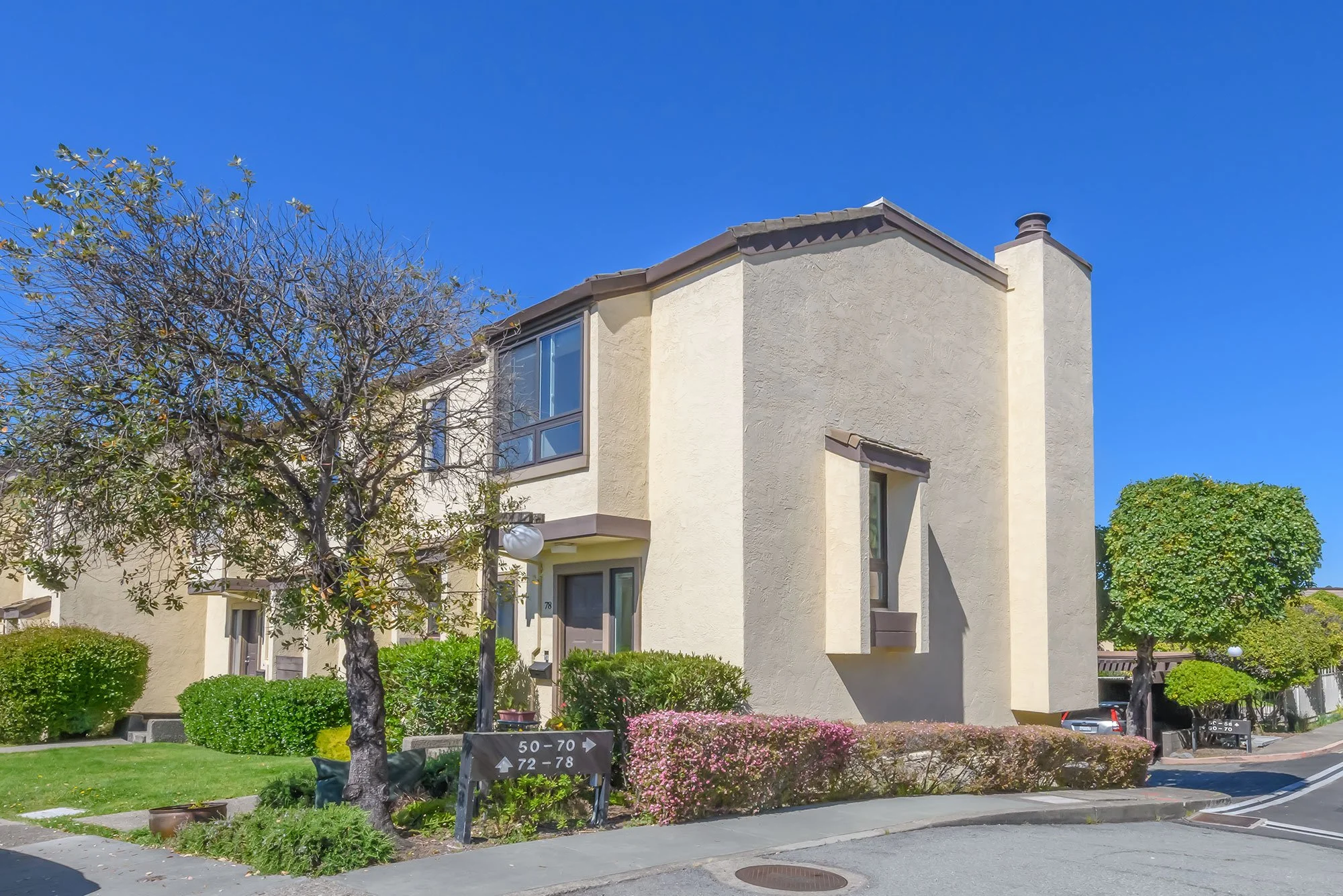 A beige two-story apartment building with a sloped roof and several windows, situated on a street corner with trees, shrubs, and flowers in front. There are street address signs and a parking area visible.