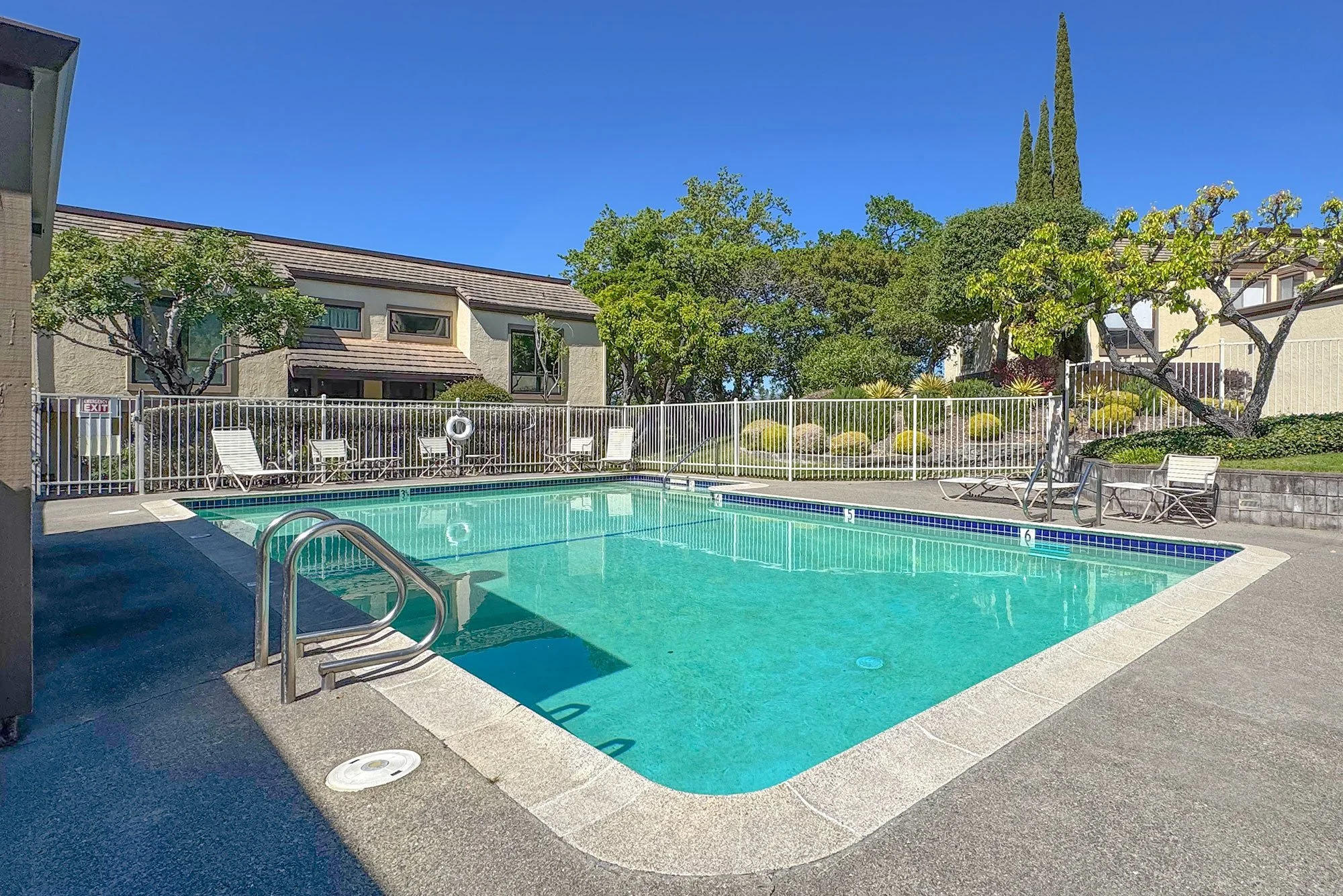 Empty swimming pool on a sunny day, surrounded by lounge chairs, trees, plants, and residential buildings.