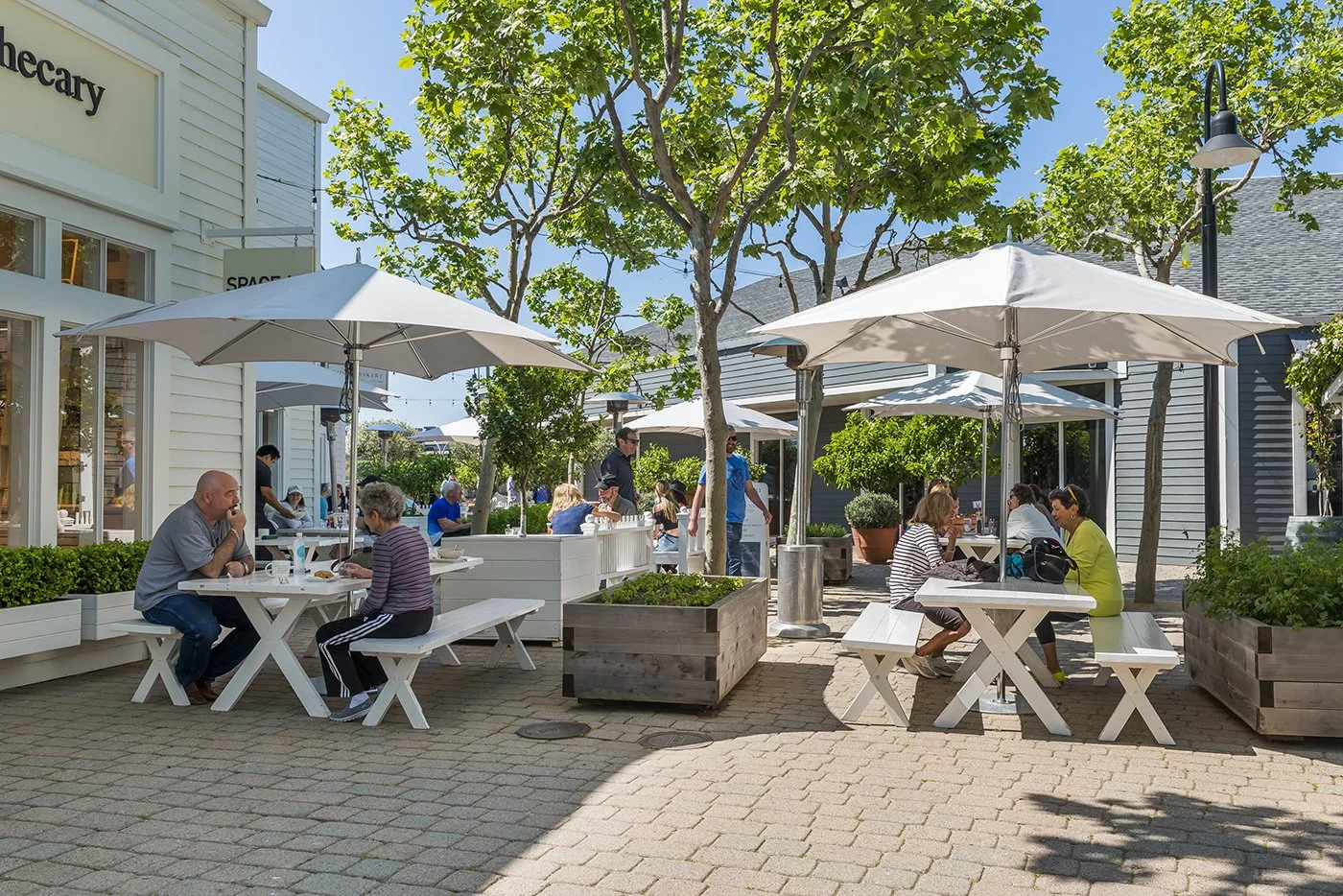 People dining outdoors at a shopping plaza or outdoor mall with white tables, umbrellas, and trees under a clear blue sky.