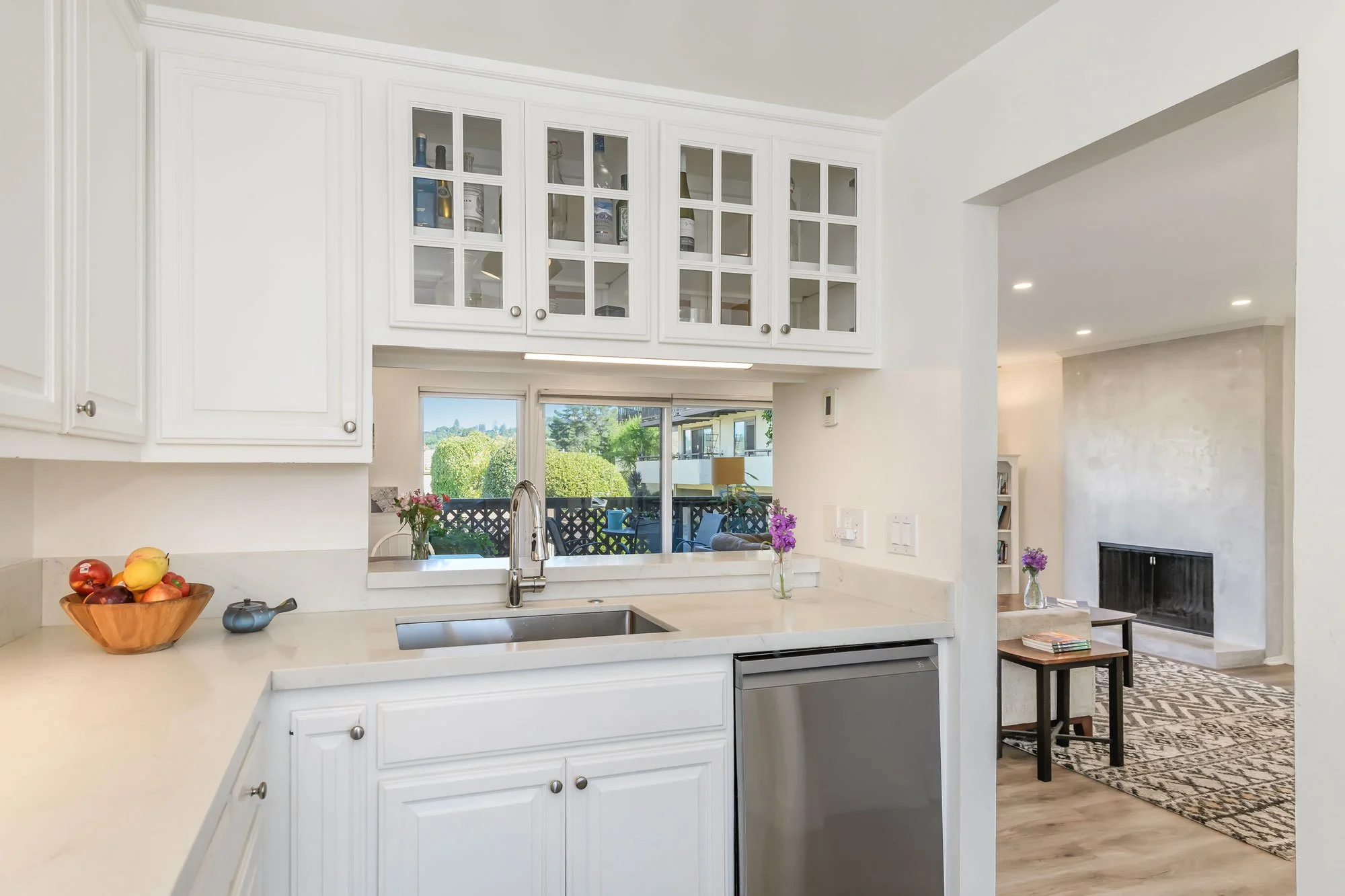 White kitchen with upper glass-front cabinets, a window above the sink, and a view into a living room with a fireplace and area rug.