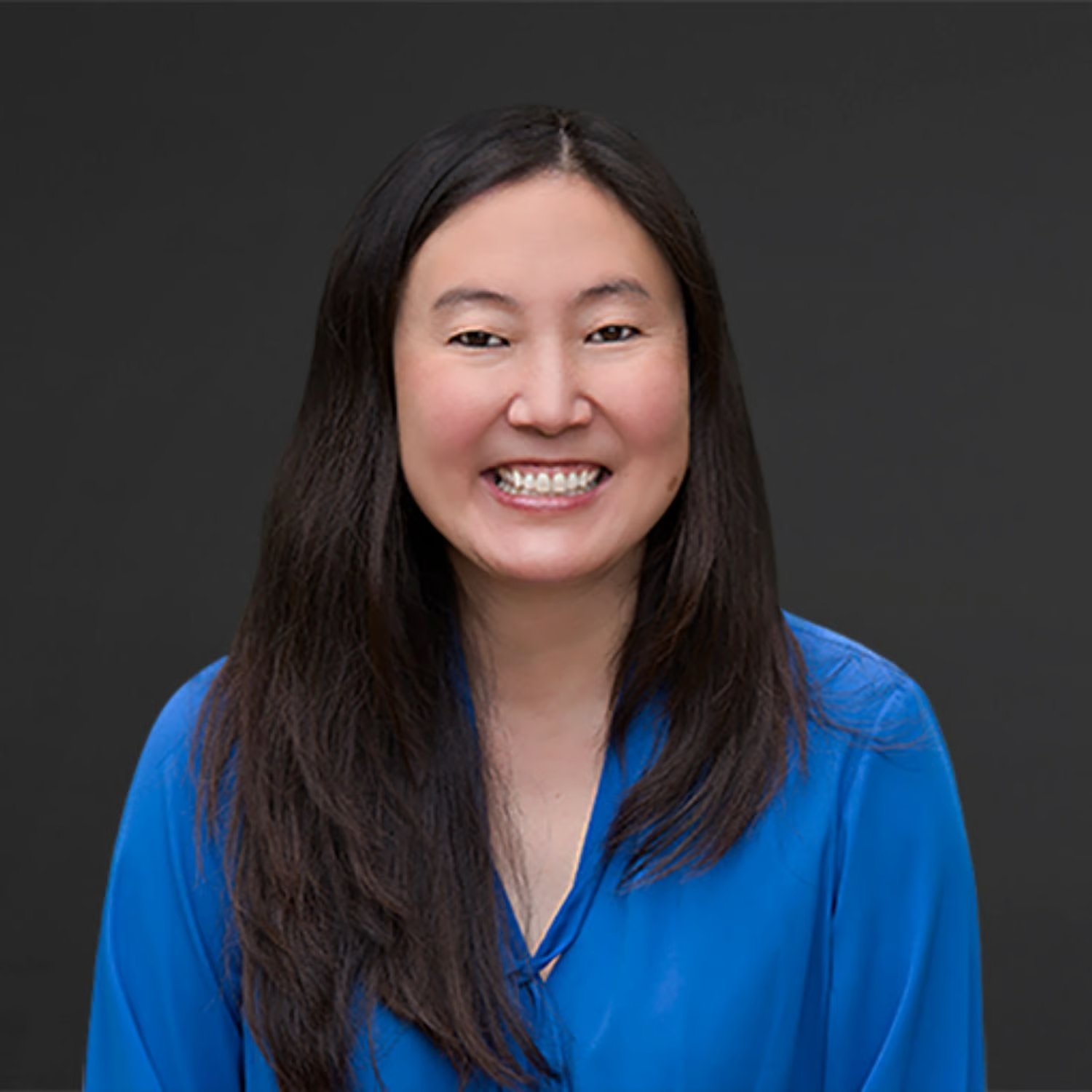 Portrait of a smiling woman with long dark hair, wearing a blue top, against a dark gray background.