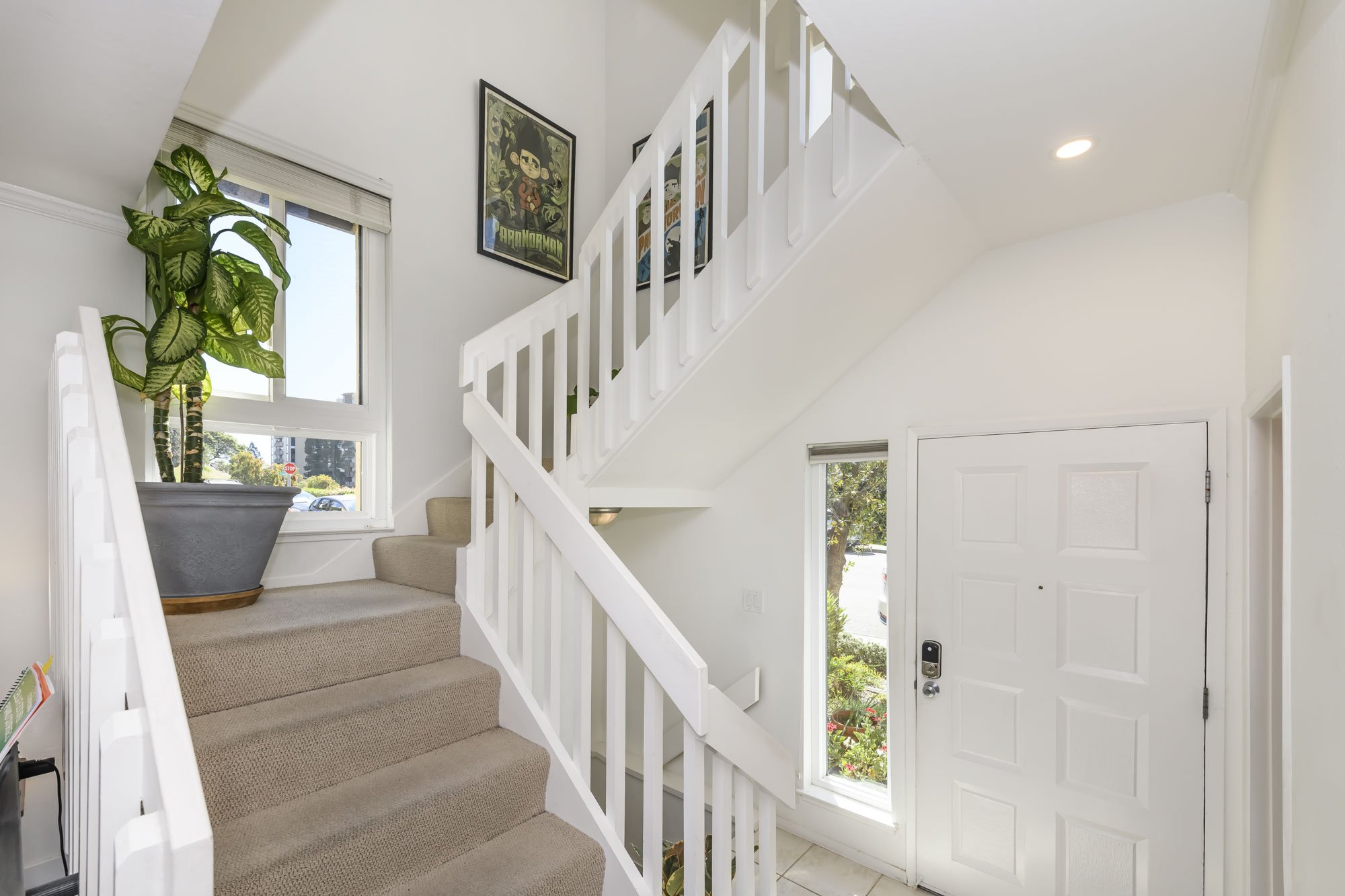 Bright entryway with carpeted stairs, a white front door with a digital lock, a small window, decorative pictures on the wall, and a large potted plant by the window.
