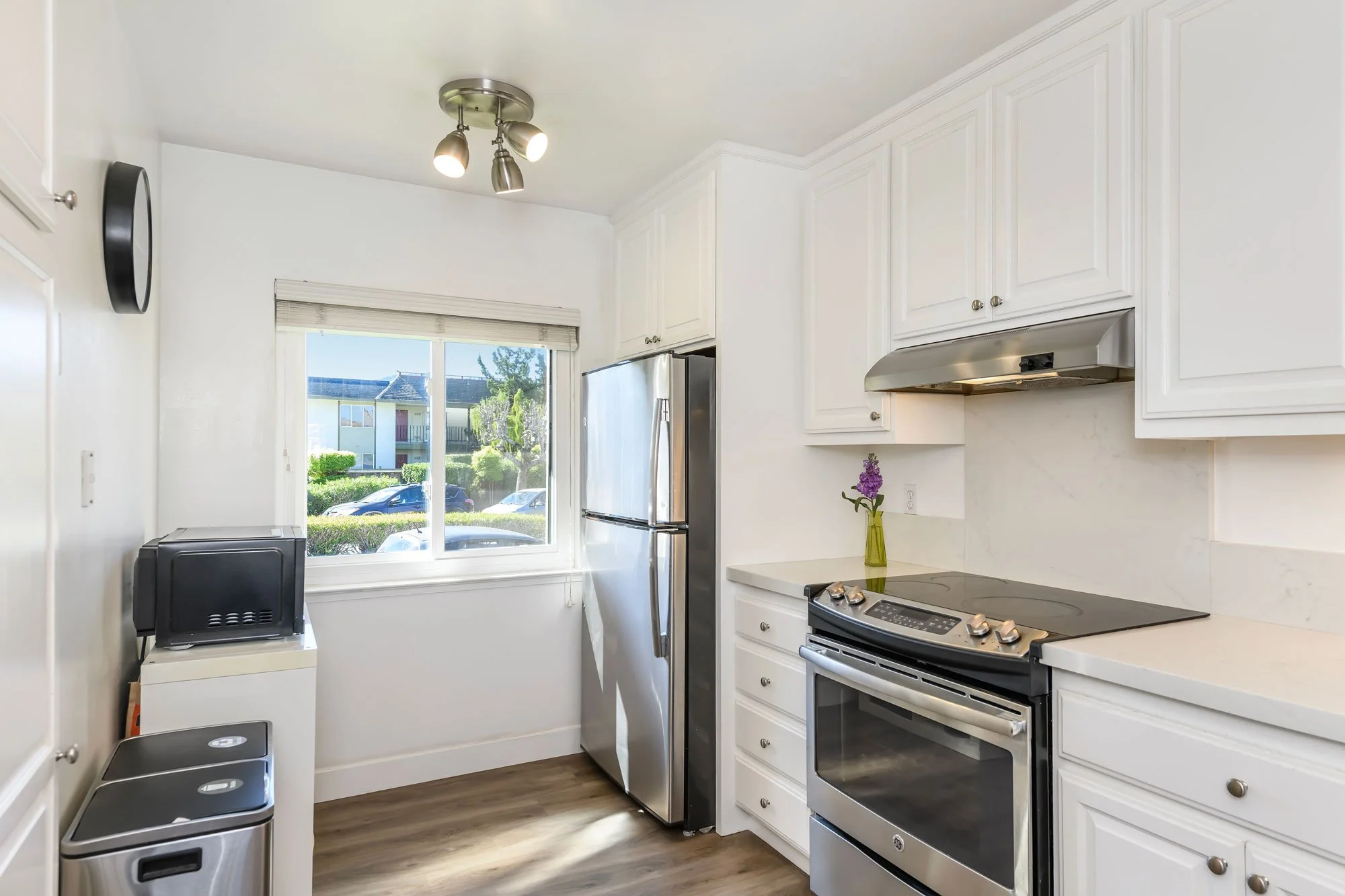 Kitchen with white cabinets, stainless steel refrigerator, stove, and a window showing an outside view of parked cars and greenery.
