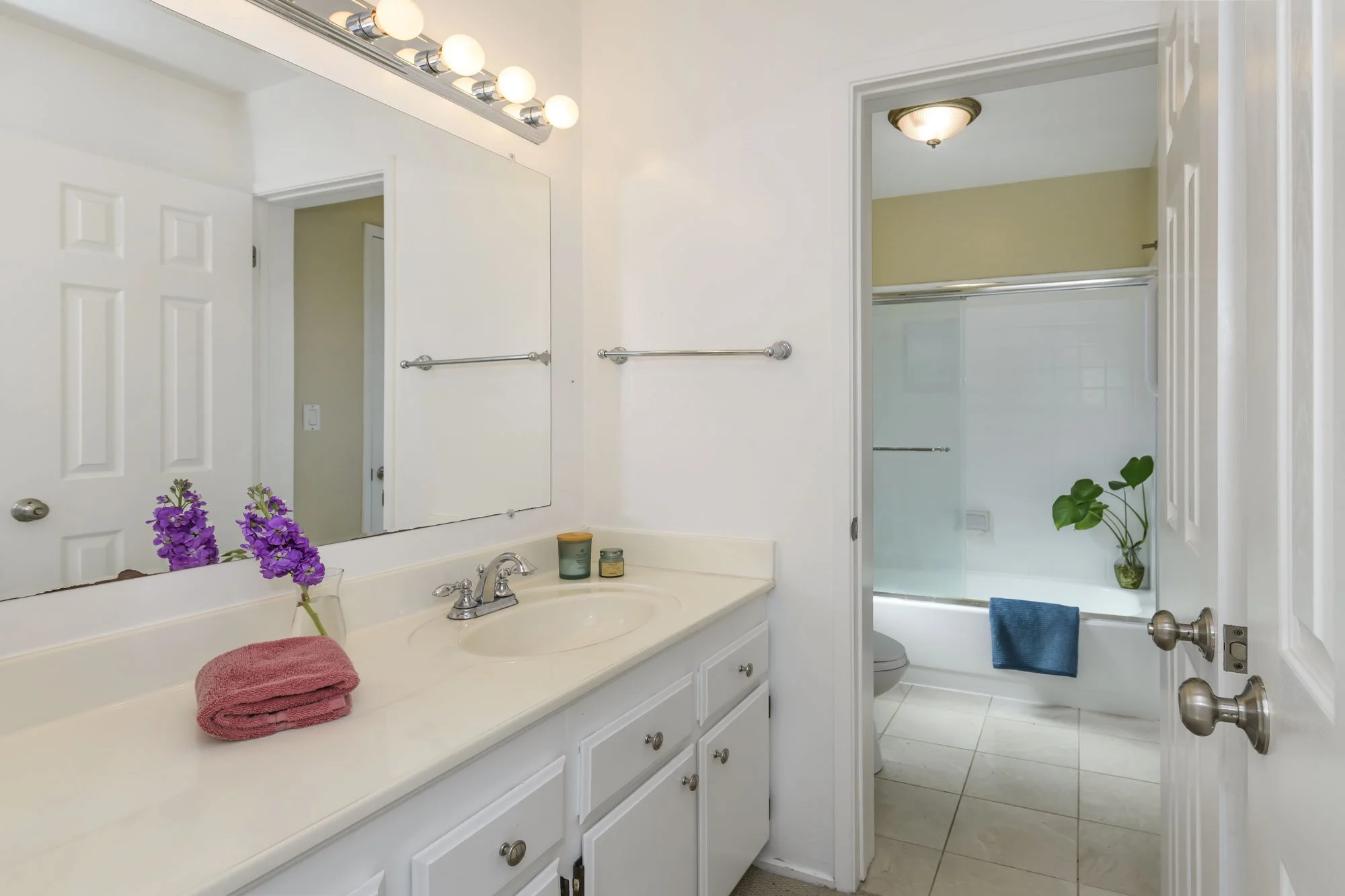 Bathroom with white vanity, mirror, and purple flowers on the countertop. Visible shower and toilet through open door. Towel rack and a hanging blue towel.