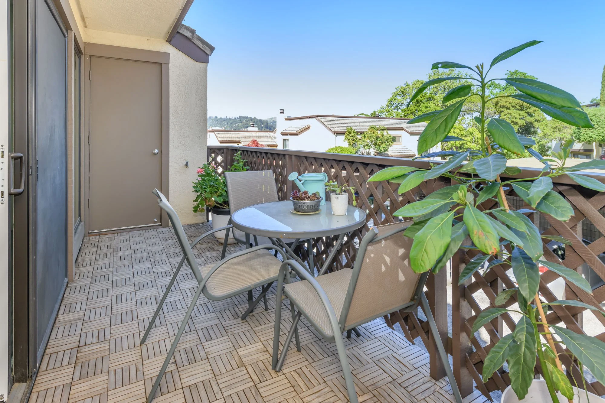 A small outdoor balcony with a round table, four chairs, and potted plants under a clear blue sky.