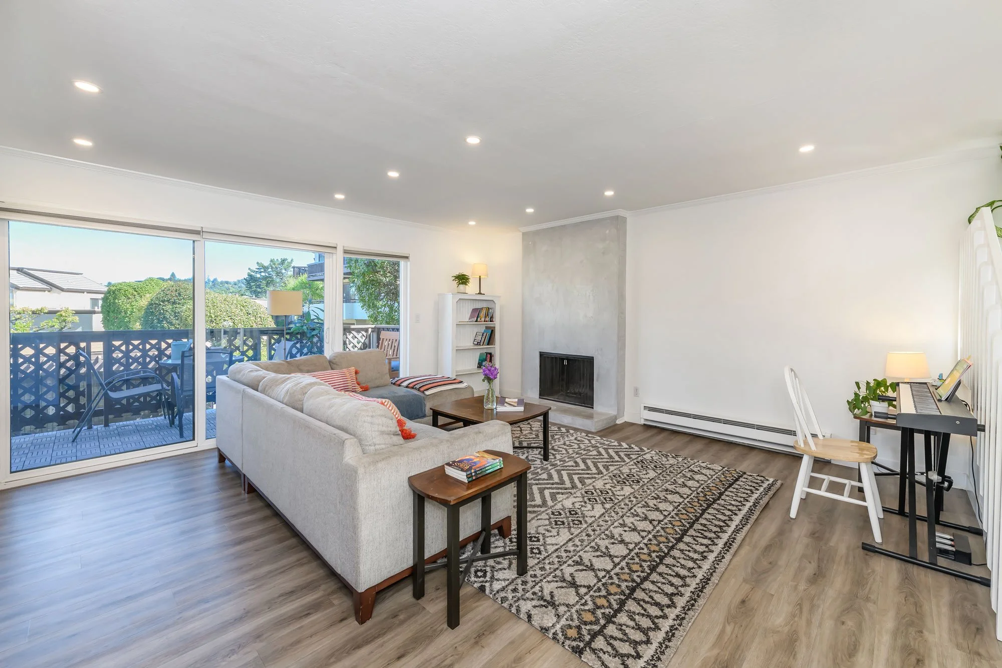 Living room with a white sectional sofa, a coffee table with flowers, a rug, a fireplace, a bookshelf, and a sliding glass door leading to a balcony with outdoor furniture. Bright, modern, and minimalistic decor.