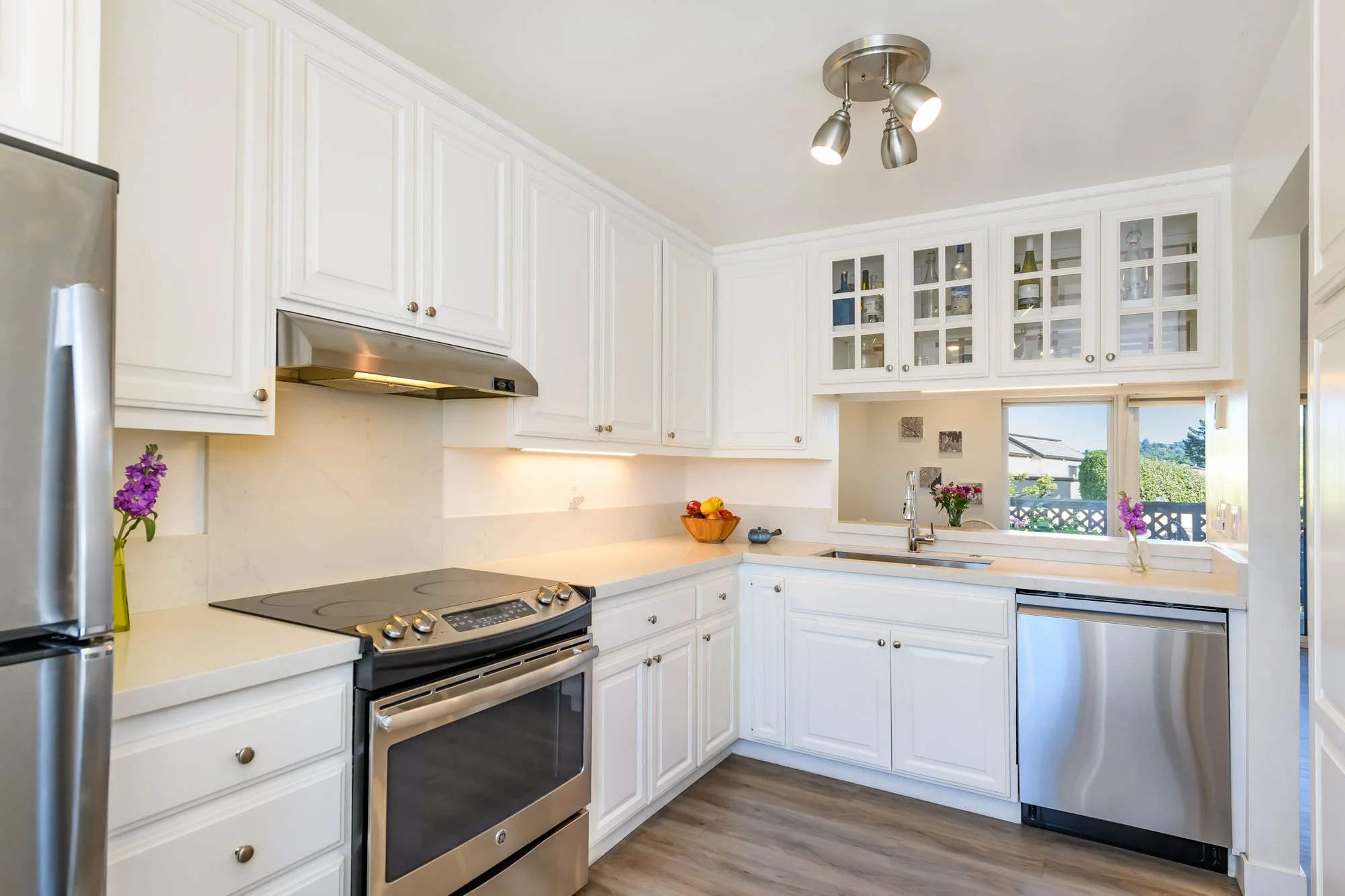 White kitchen with stainless steel appliances, white cabinets, a small window above the sink, and decorative purple flowers on the counter.