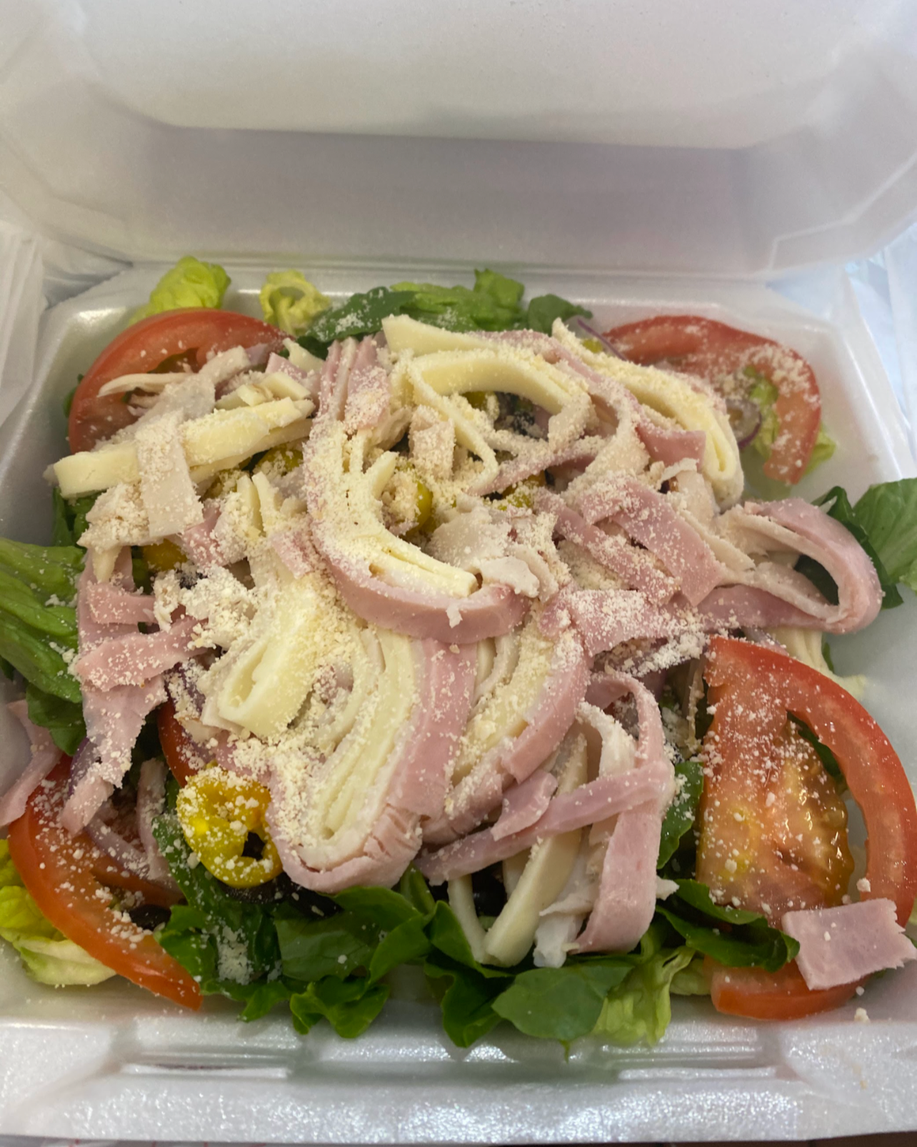 Mixed salad with sliced tomatoes, shredded cheese, ham, lettuce, and green peppers in a foam takeout container