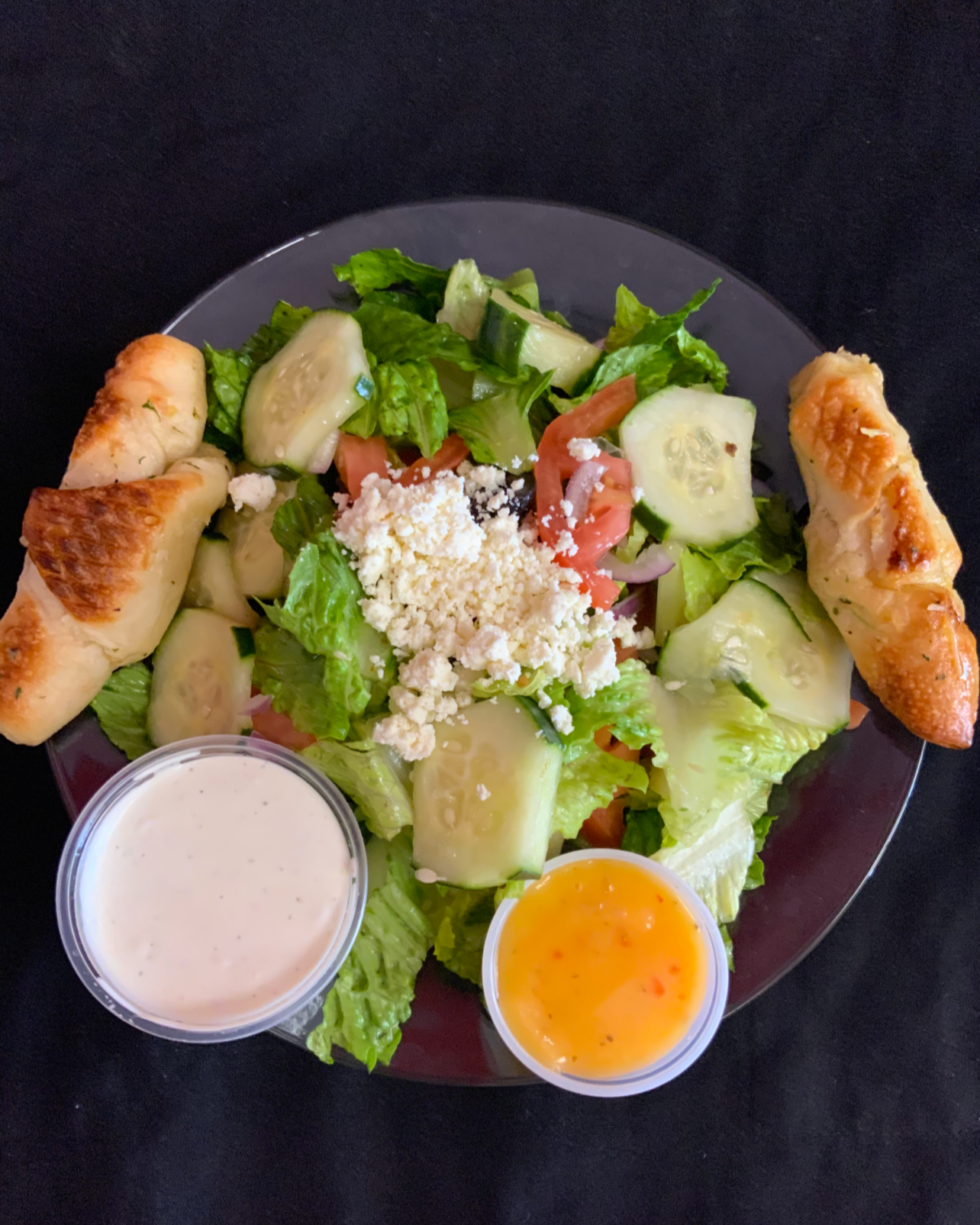 Salad with lettuce, cucumbers, tomatoes, cheese, two bread rolls, and two small cups of dressing, one creamy and one with an orange hue.