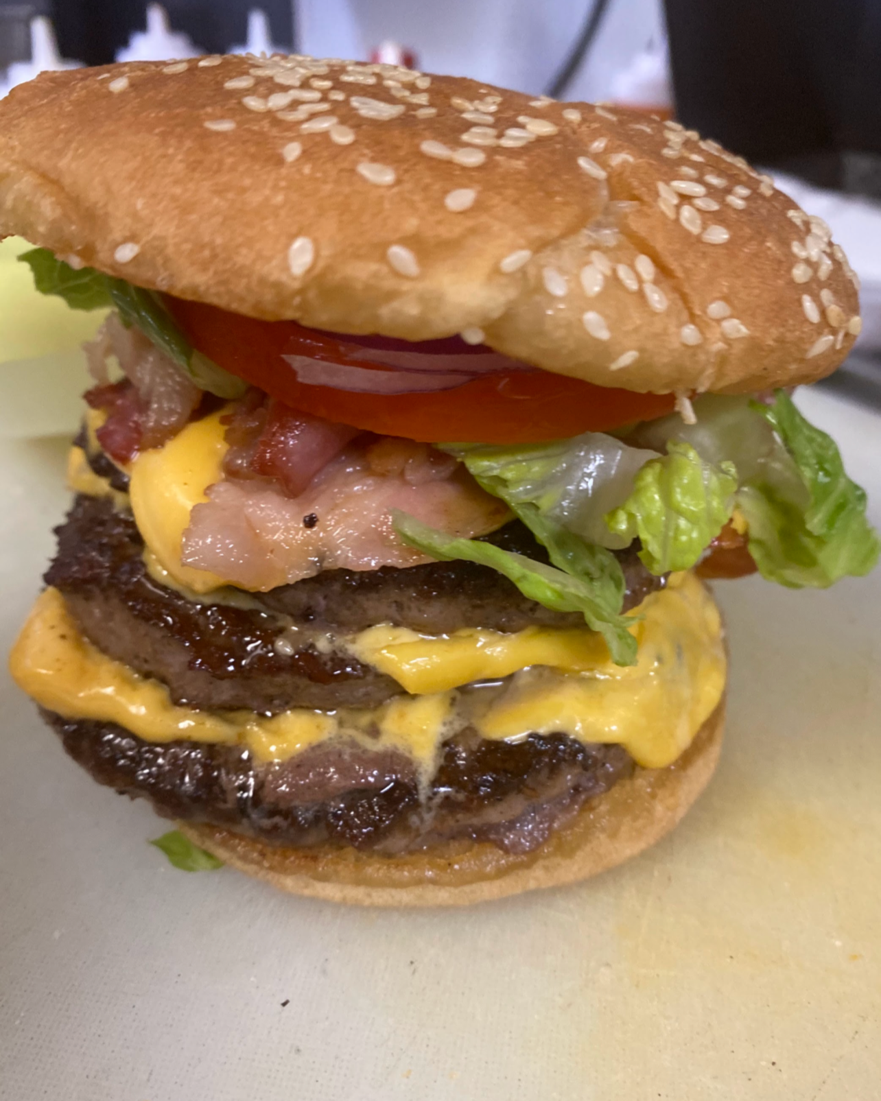 Close-up of a cheeseburger with sesame seed bun, lettuce, tomato, bacon, cheeseburger patties, scrambled eggs, and melted cheese.
