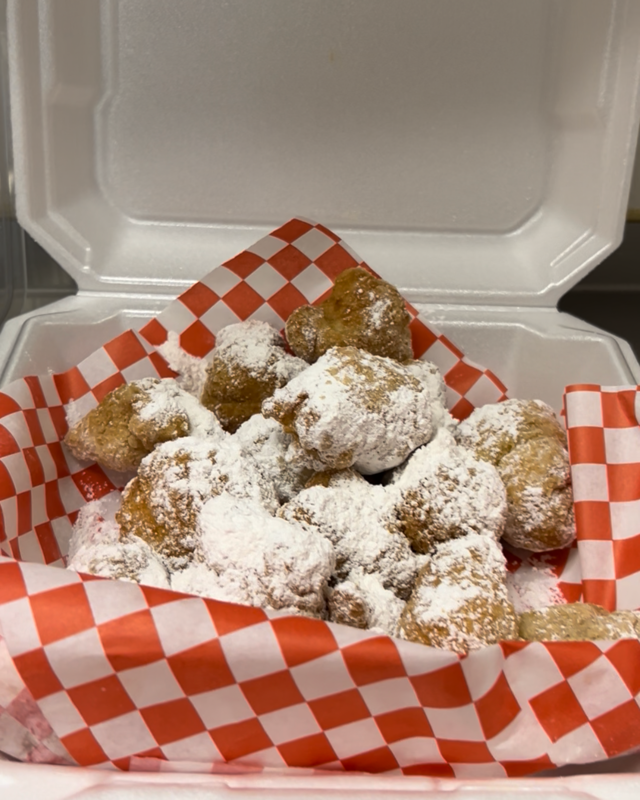 Fried dough balls dusted with powdered sugar in a red and white checkered-lined container
