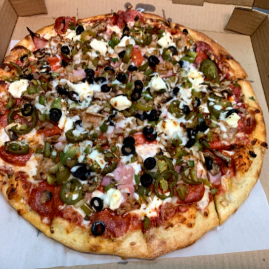 A large pizza with toppings including black olives, green peppers, red onions, pepperoni, ham, and cheese in a cardboard delivery box.