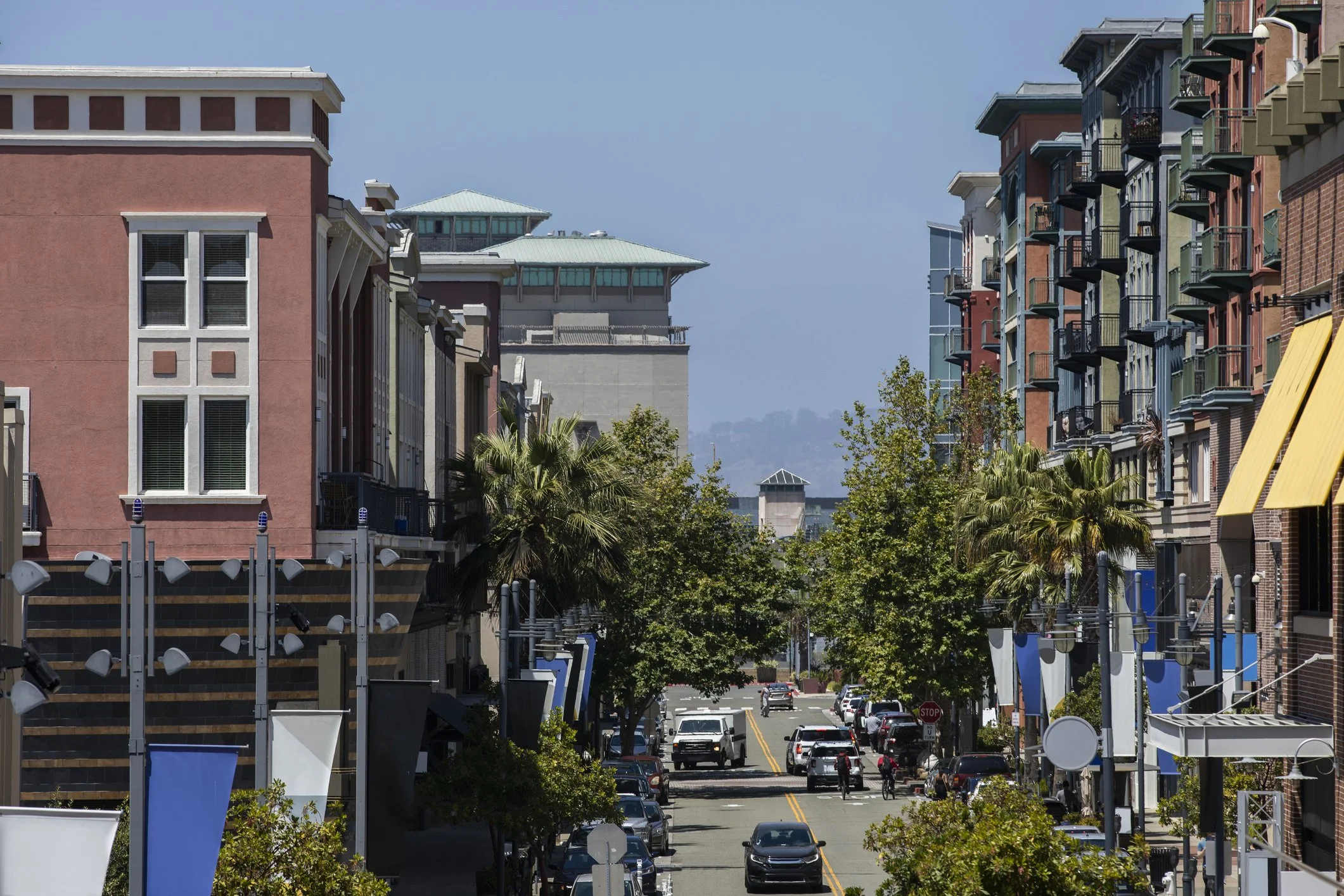 A city street lined with colorful multi-story buildings, with parked cars and trees along the sidewalk, under a clear blue sky.