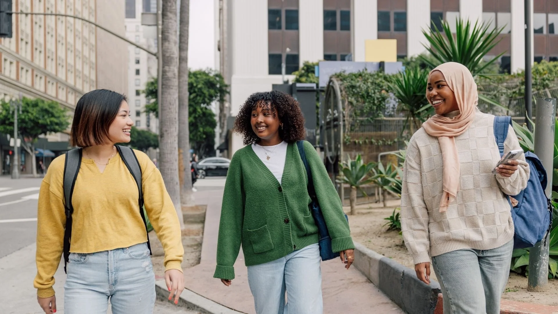 Three women walking on city sidewalk, smiling and talking, with tall buildings, trees, and plants in the background.