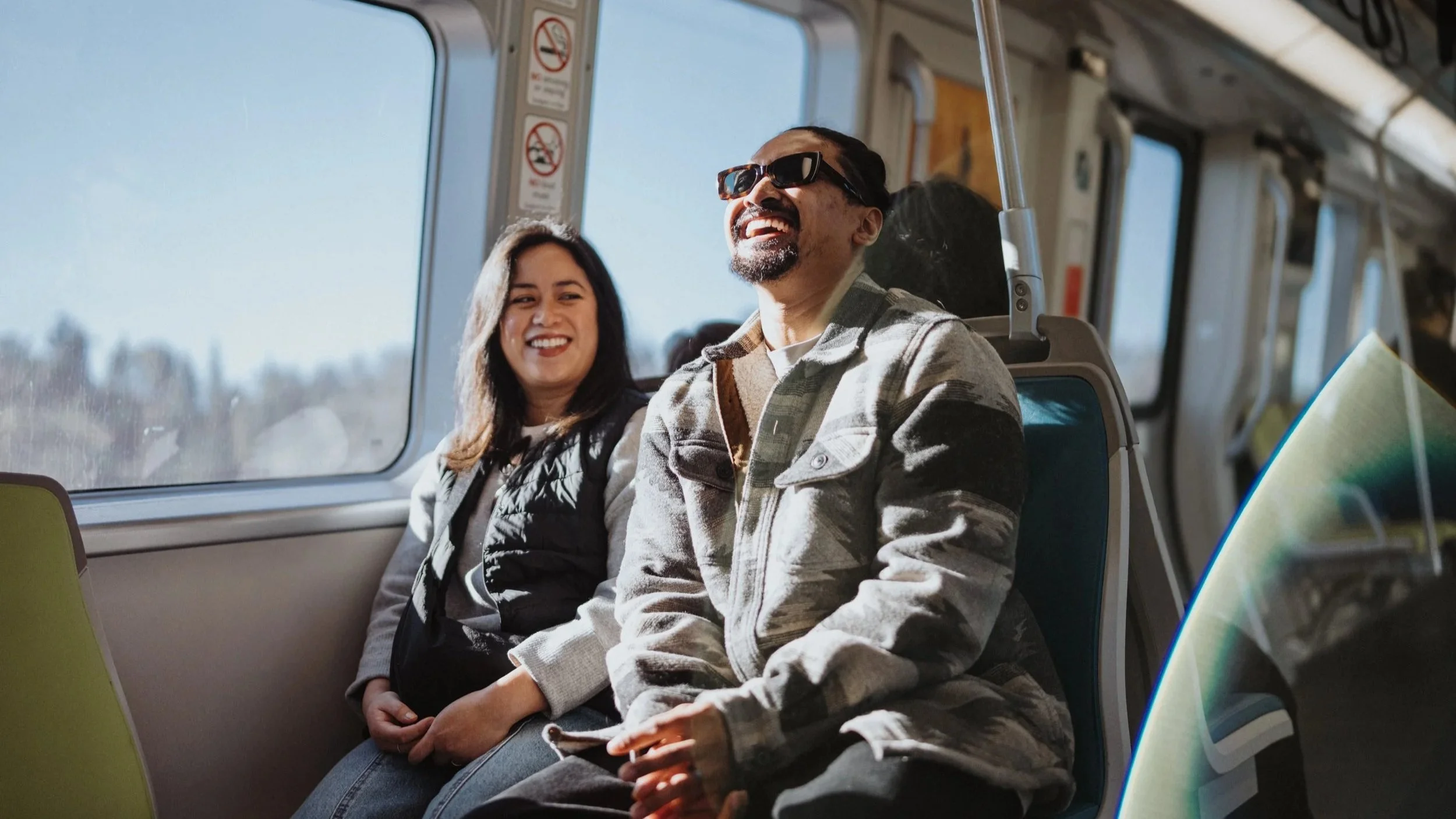 Two people sitting on a bus, smiling and laughing, with sunlight streaming through the windows.