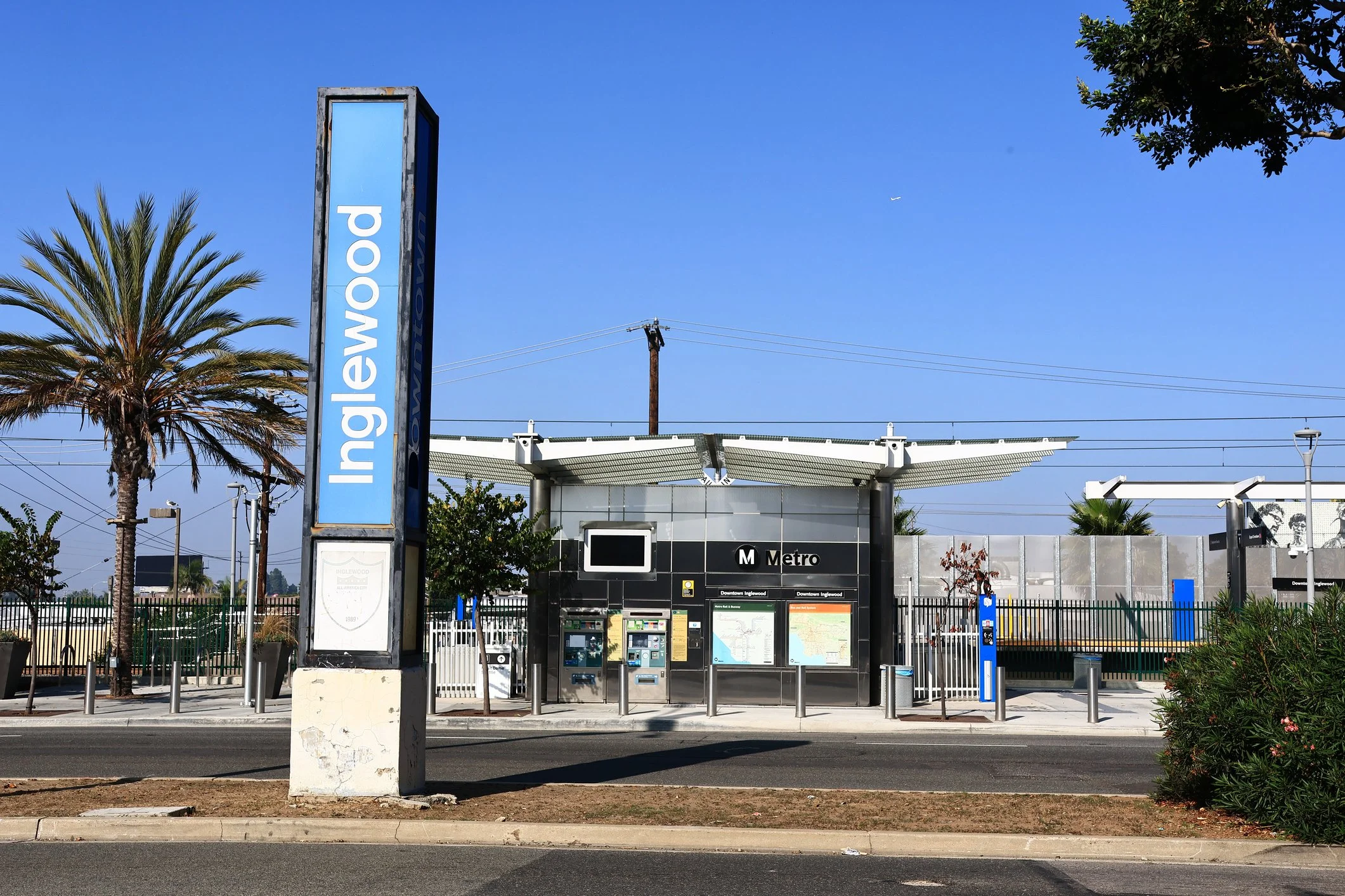 Inglewood Metro station entrance with a large vertical sign, ticket machines, map displays, surrounded by palm trees, clear blue sky, and fencing.