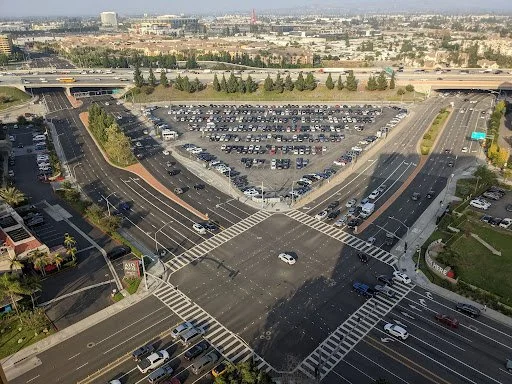 An aerial view of a large parking lot and intersection in an urban area. The parking lot is filled with many cars, and the intersection has multiple lanes with a few vehicles.