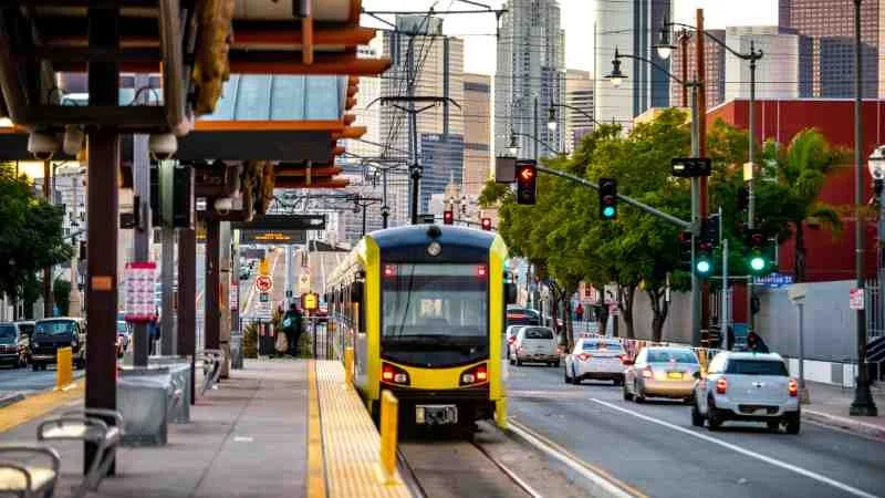 City street scene with a yellow and black light rail train approaching a station, surrounded by cars, trees, and city skyscrapers.