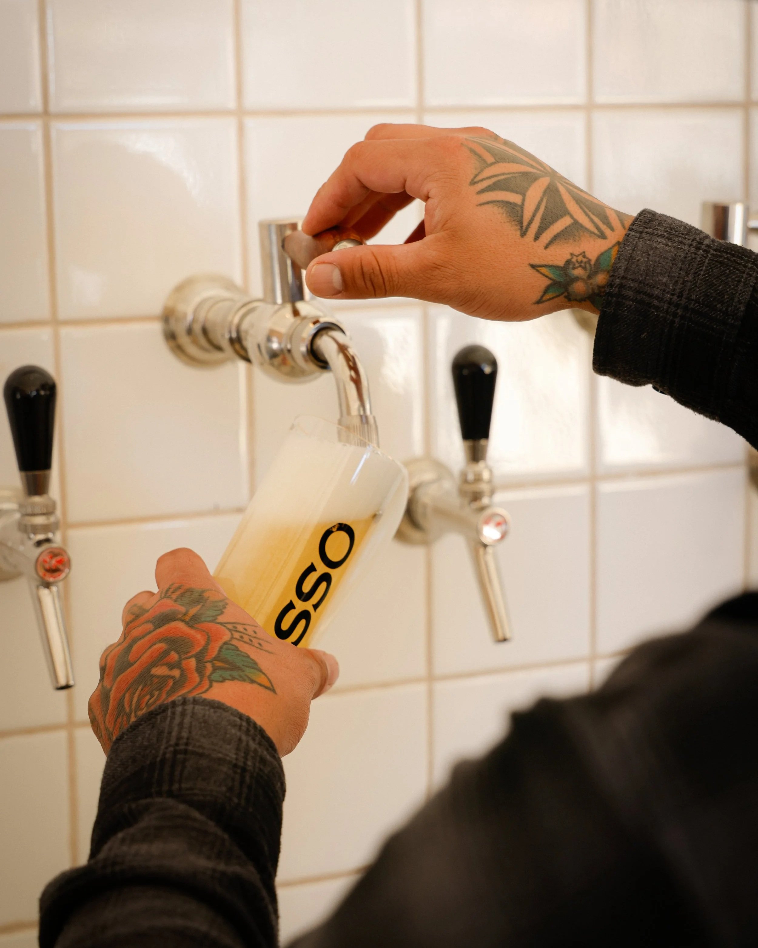 Person with tattoos pouring beer from tap into a glass marked with the word 'OSSO' in a setting with tiled wall and multiple beer taps.