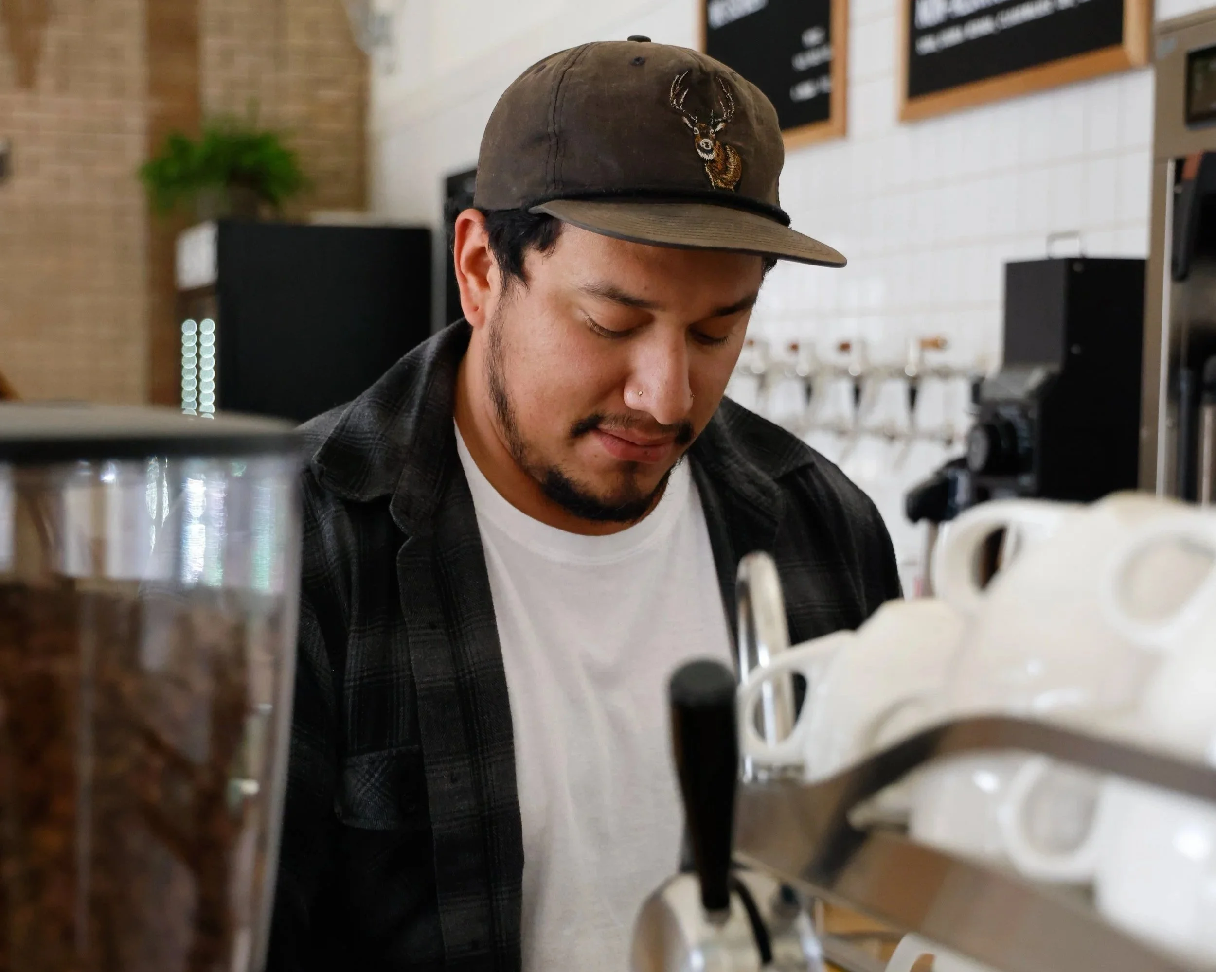 A man with dark hair, beard, and a nose piercing using an espresso machine in a coffee shop.