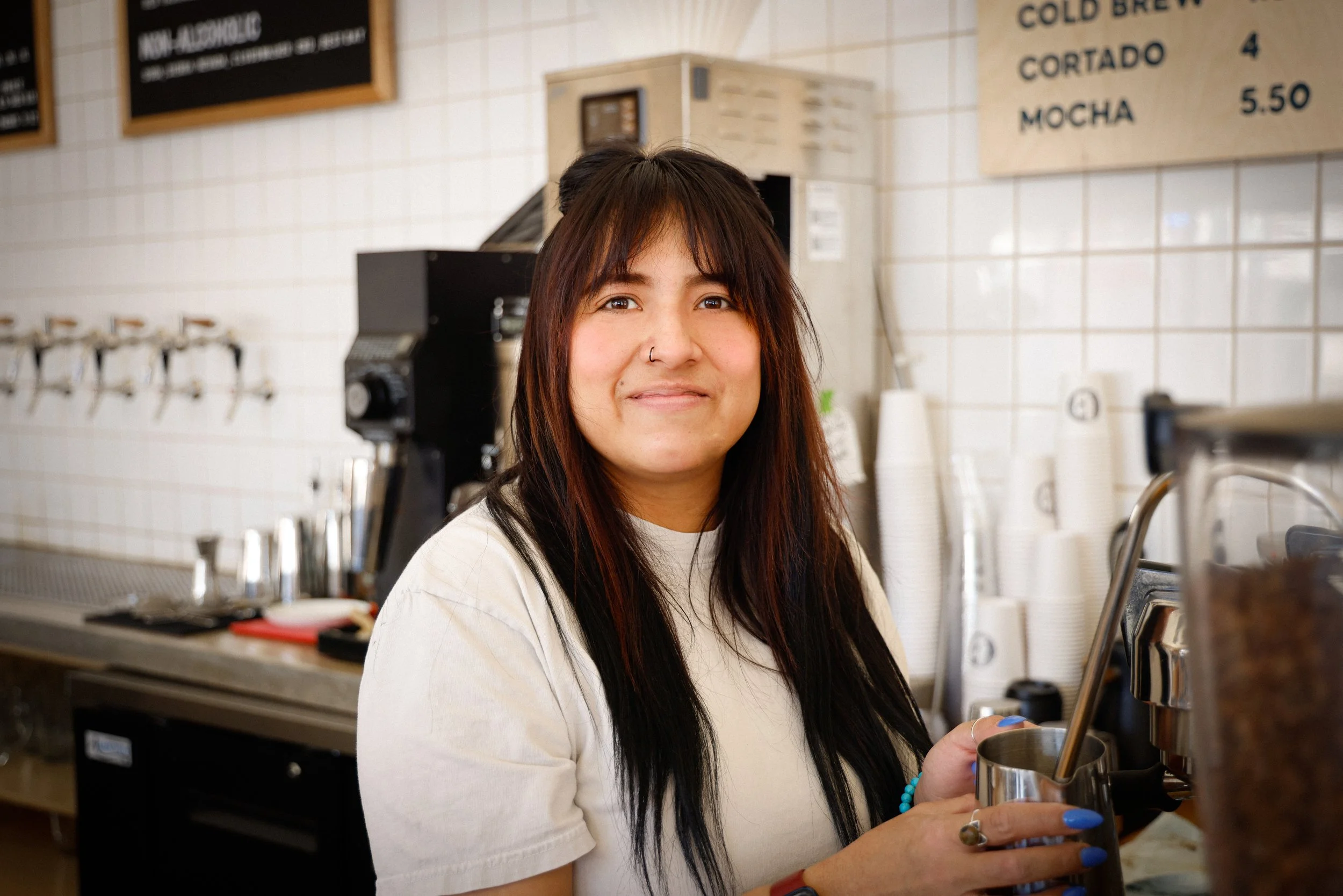 A woman with brown and black hair, wearing a white t-shirt, smiling behind a coffee machine in a cafe, holding a metal pitcher.
