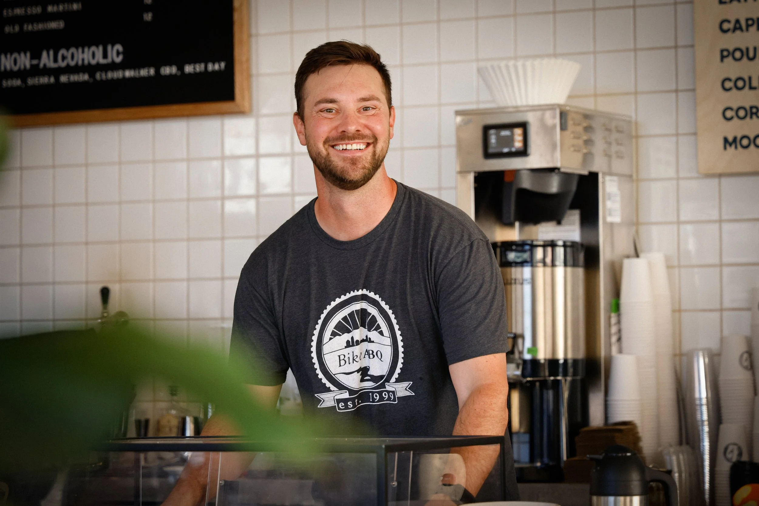 A woman with dark hair and glasses drinks iced coffee from a glass mug, sitting across from another person at a cafe, with 'LASSO' written on a sign in the background.