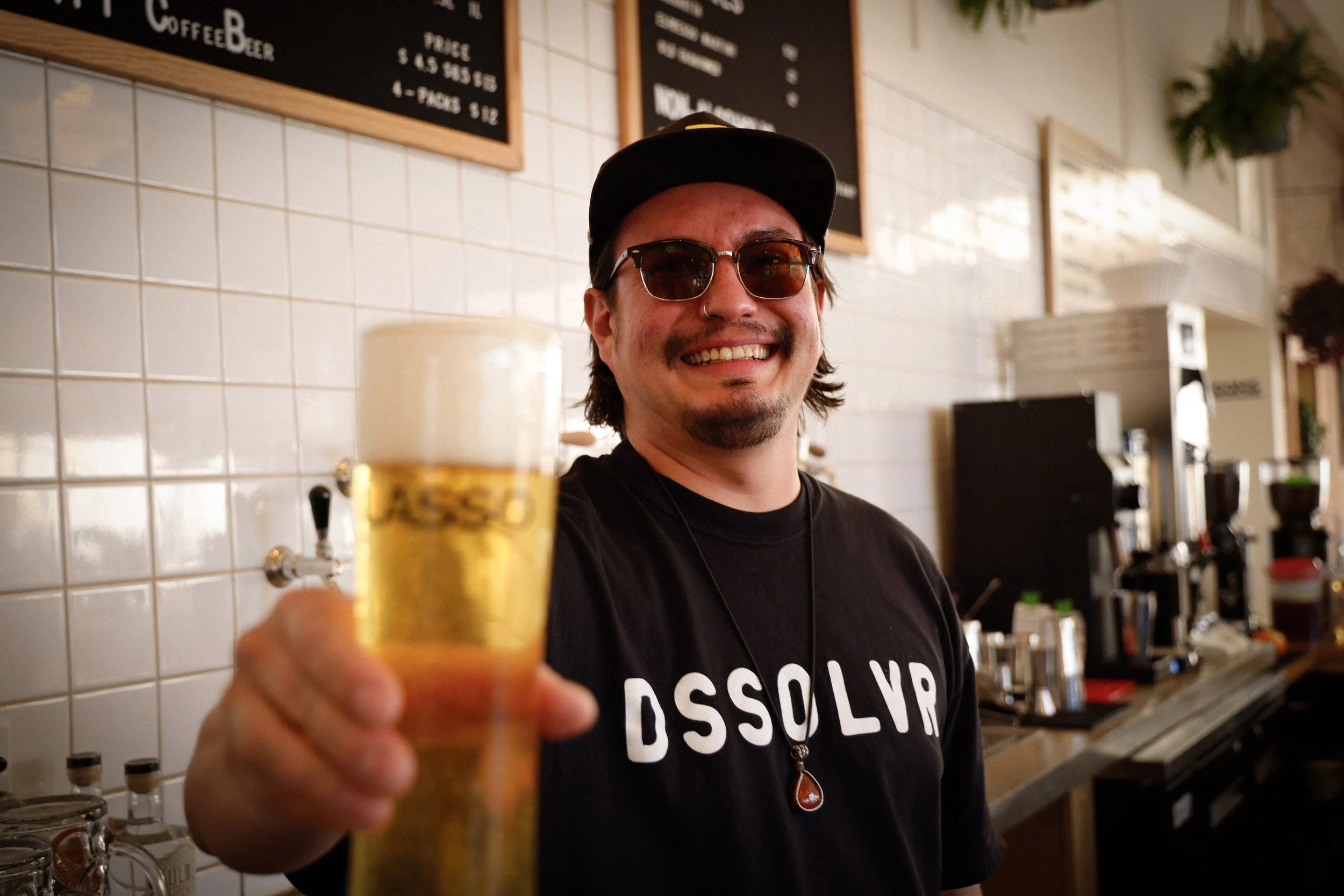A smiling man with sunglasses and a black cap holding a glass of beer in a bar or cafe setting.