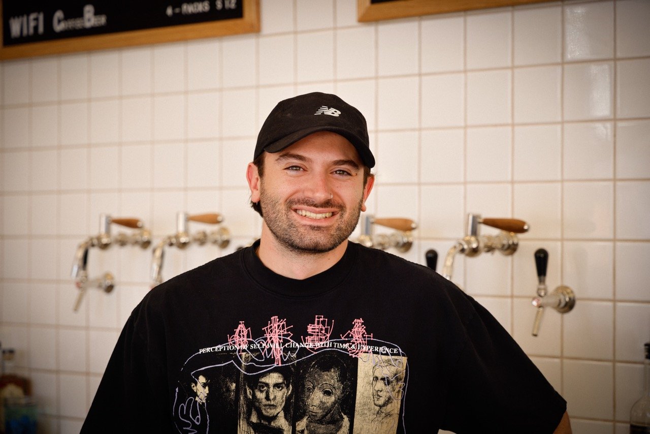 A smiling man wearing a black cap and a black graphic T-shirt standing in front of a tiled wall with beer taps.