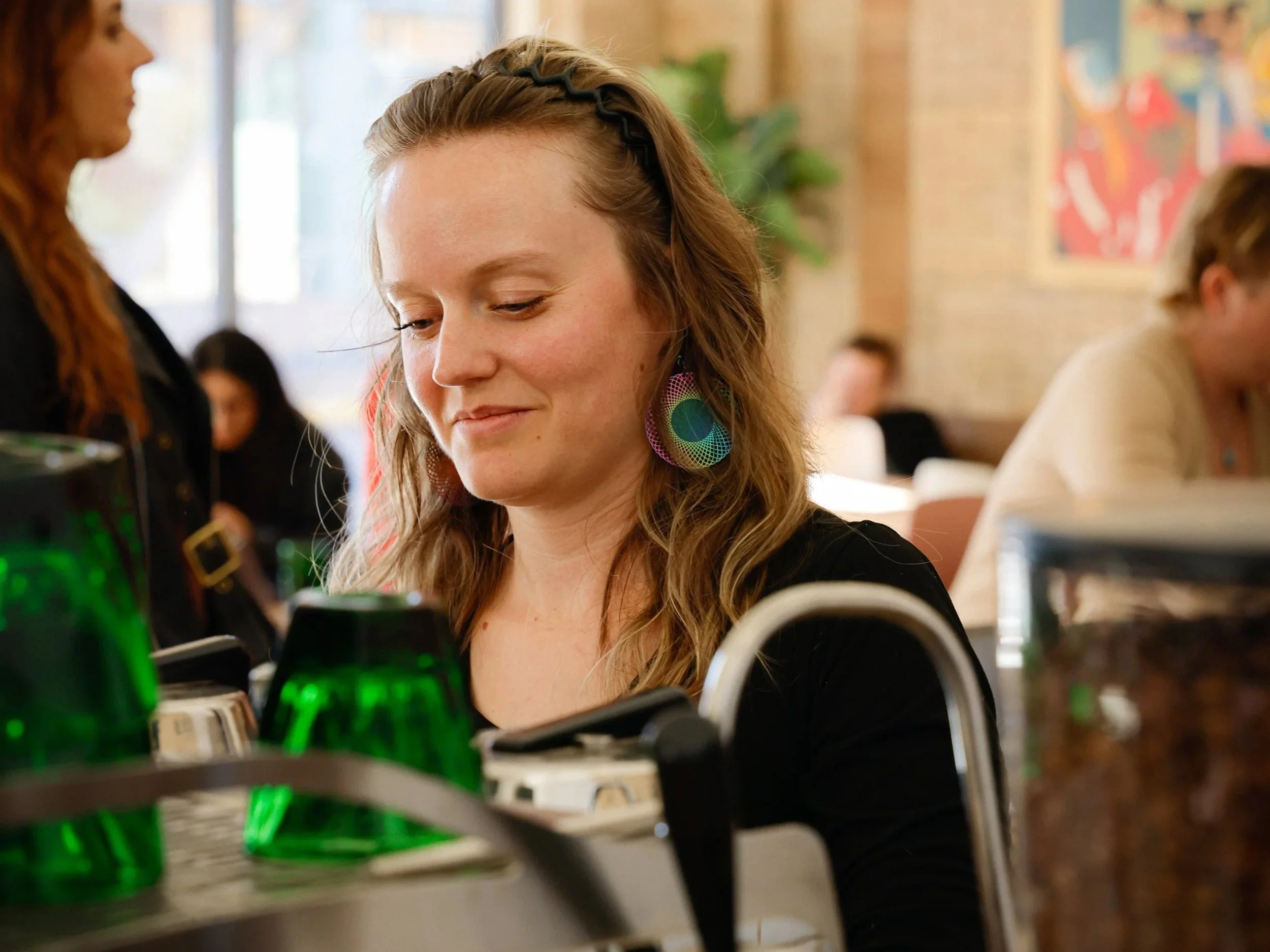 A young woman with wavy hair and colorful earrings in a cafe, smiling and looking down at a dish or object in front of her. There are other people in the background, some sitting and some standing, with a brick wall and colorful artwork on the wall.