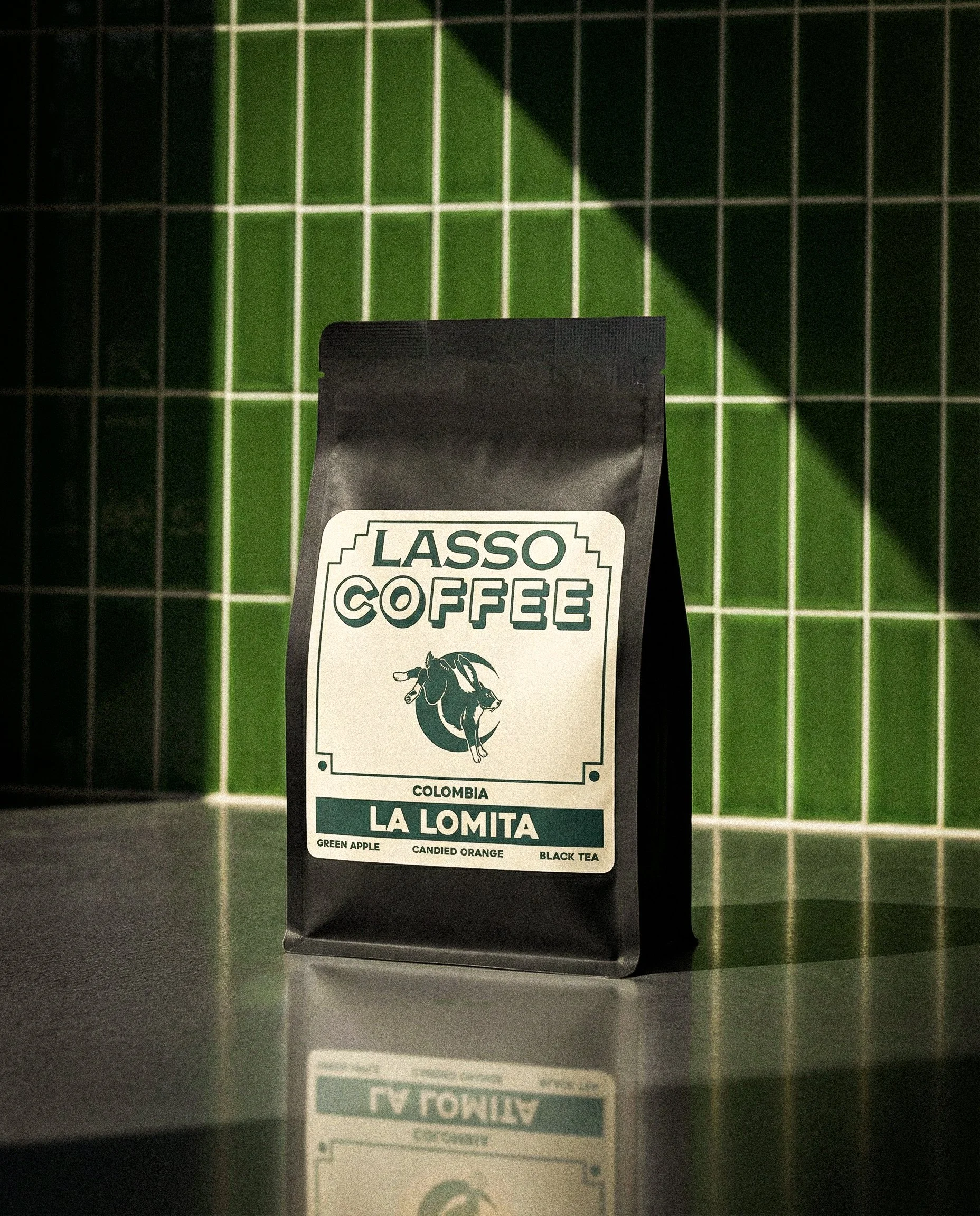 A black coffee bag labeled 'Lasso Coffee' from Colombia, with flavor notes of green apple, candied orange, and black tea, placed on a reflective surface with a green tiled wall in the background.