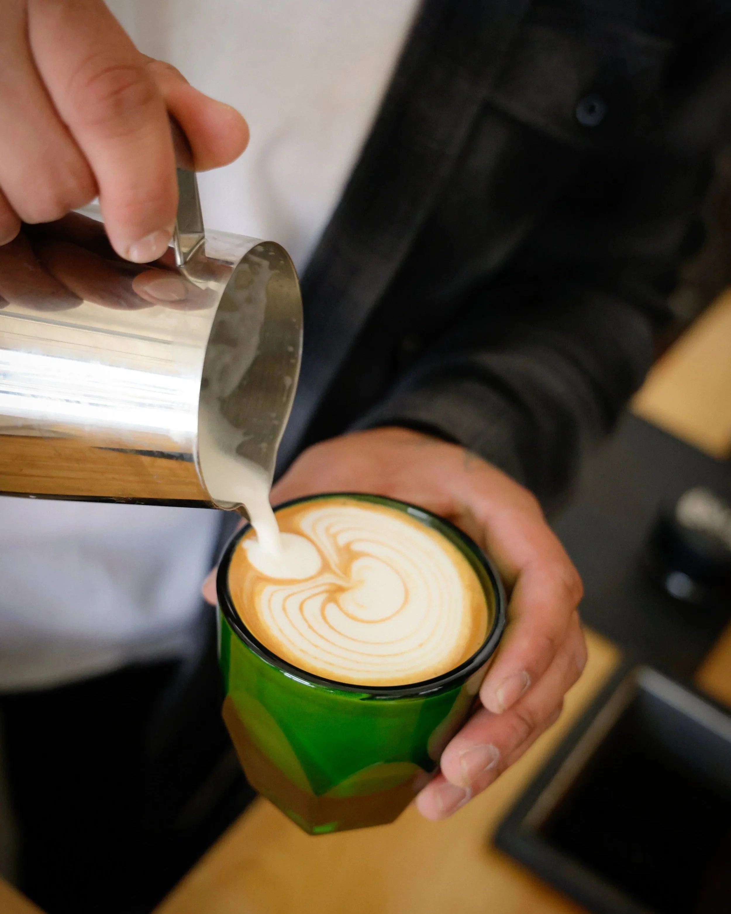 Barista pouring steamed milk into a latte with latte art in a black mug with a green and yellow design.