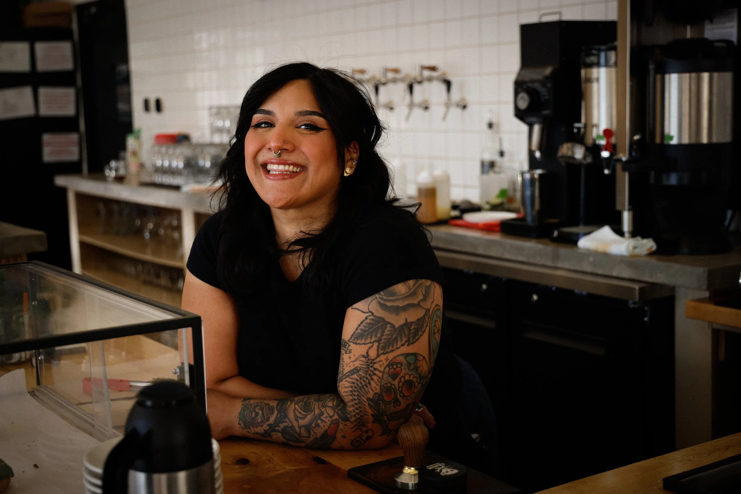 Smiling woman with black hair, tattoos on her arm, wearing a black shirt, sitting at a counter inside a cafe or restaurant.