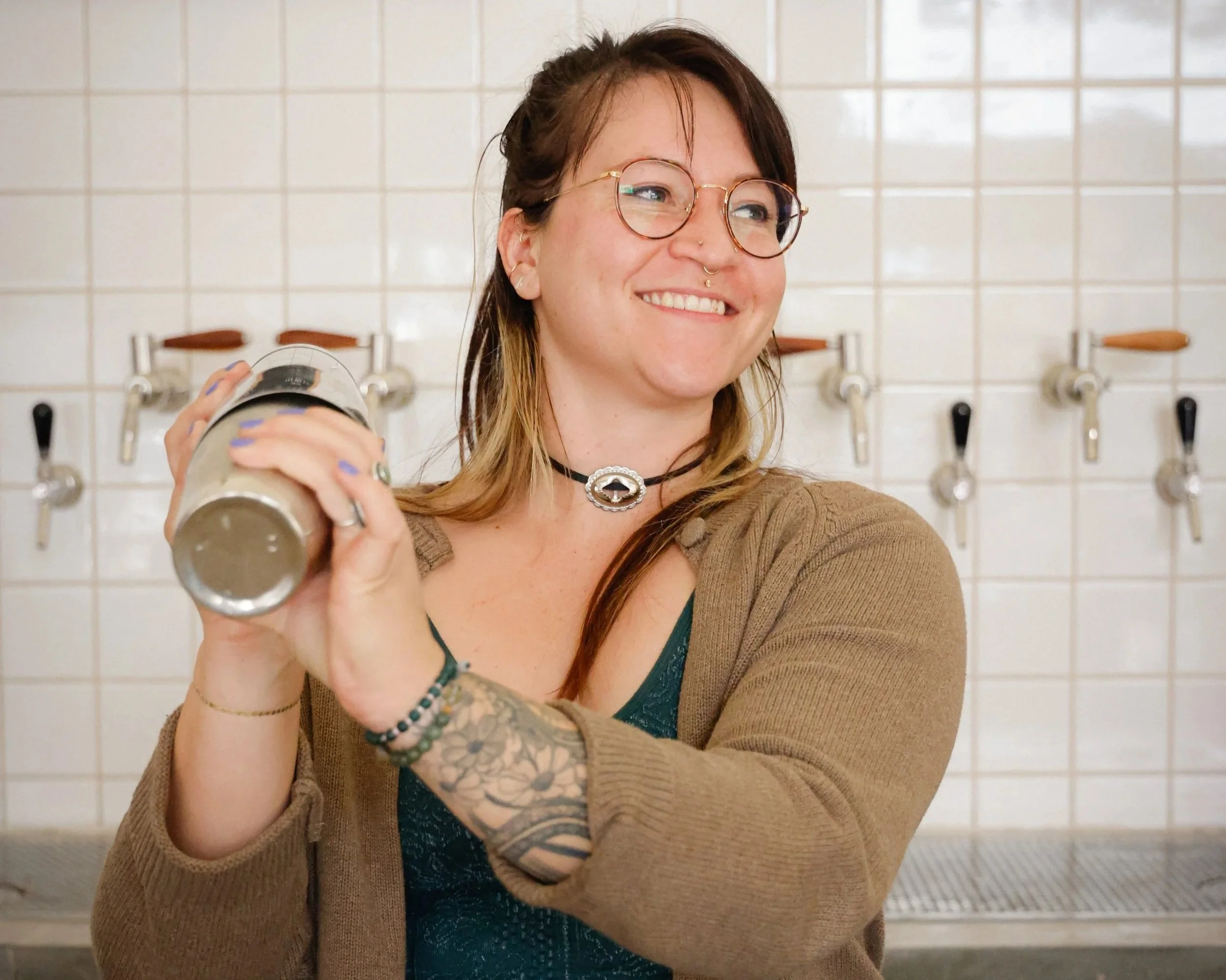 A young woman with glasses and tattoos on her arm, holding a beer stein, smiling, in a room with tiled walls and multiple beer taps in the background.