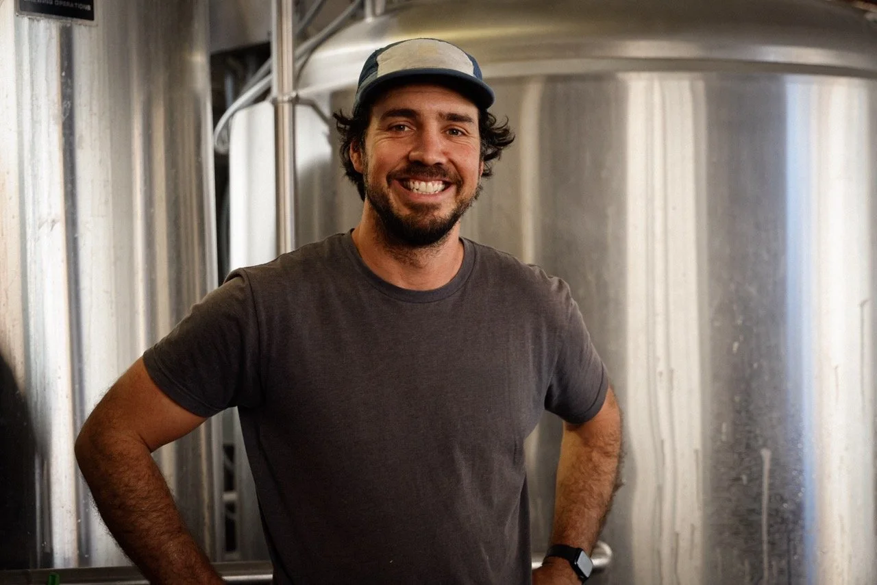 A smiling man with a beard and mustache wearing a gray t-shirt, a camouflage cap, and a wristwatch standing in front of large stainless steel tanks in a brewery or industrial setting.