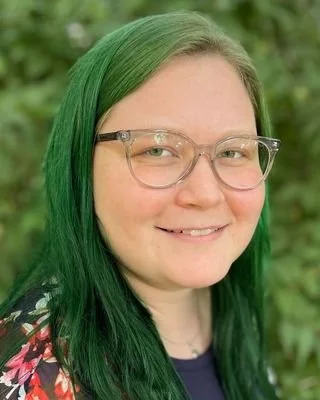 Portrait of a woman with green hair and glasses, smiling outdoors with greenery in the background.