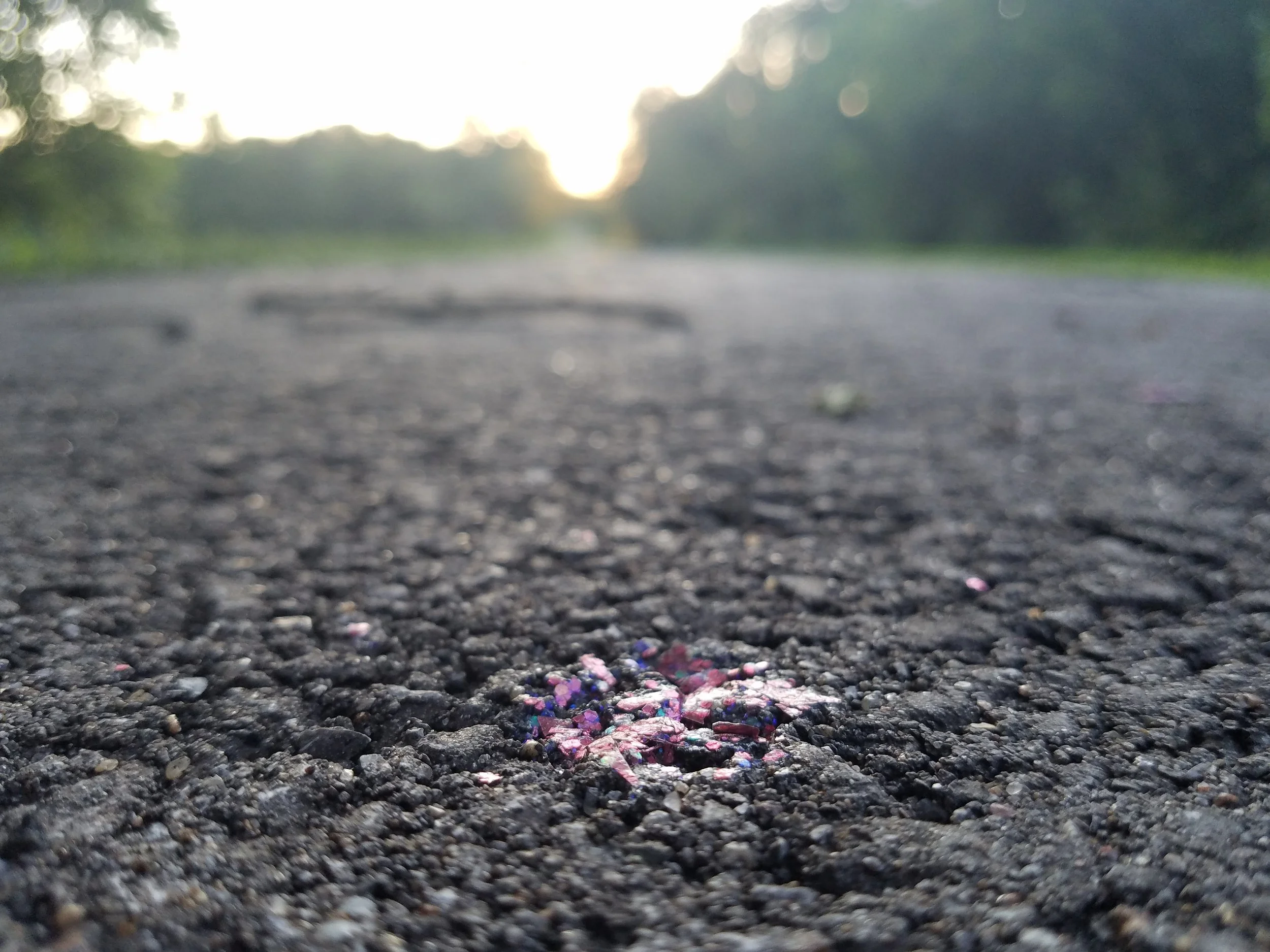 Close-up of a paved road at sunset with colorful paint splashes on the surface, surrounded by trees and greenery in the background.